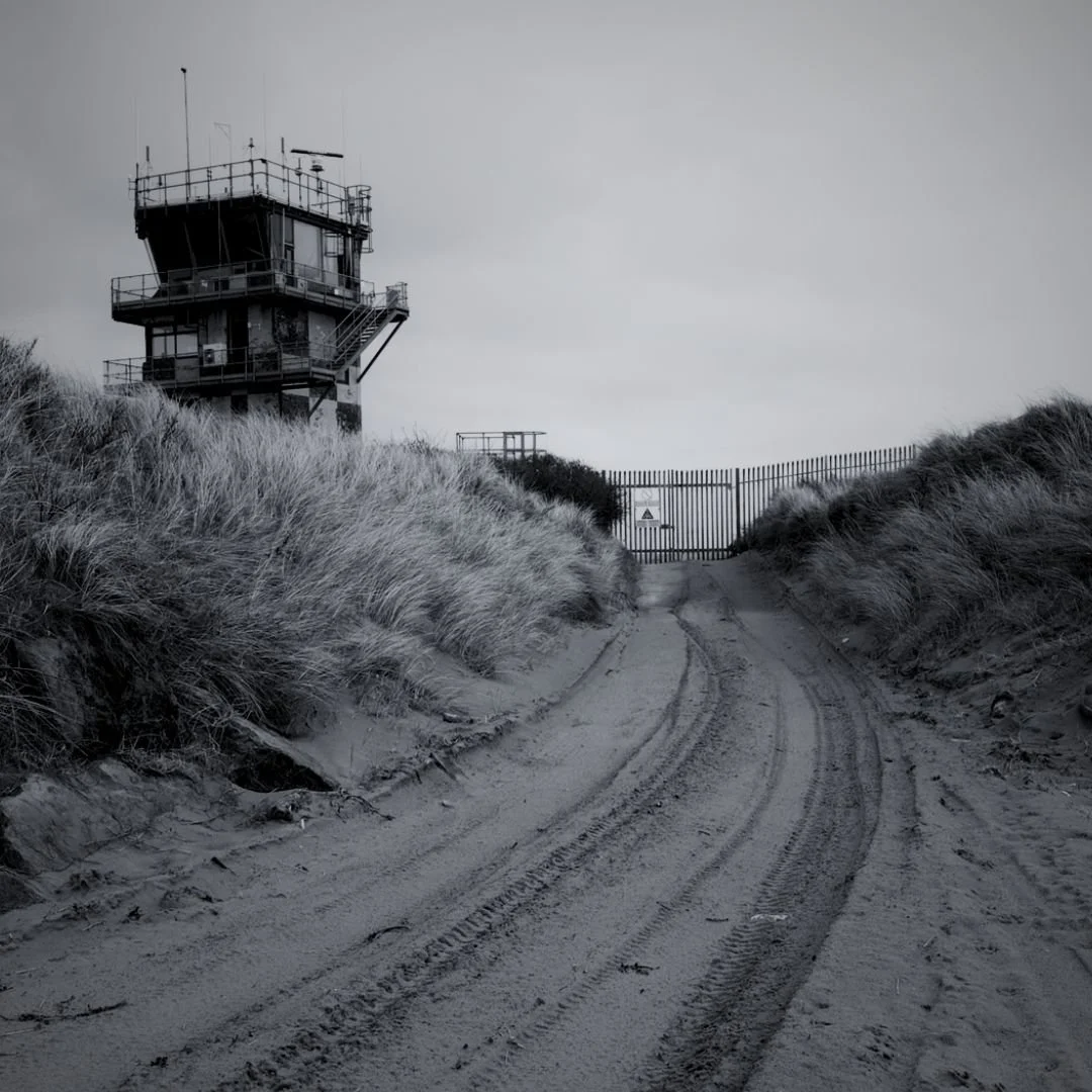 A black and white photo of a sandy pathway leading to a high guard tower and a gate, surrounded by tall sand dunes and grassy vegetation.