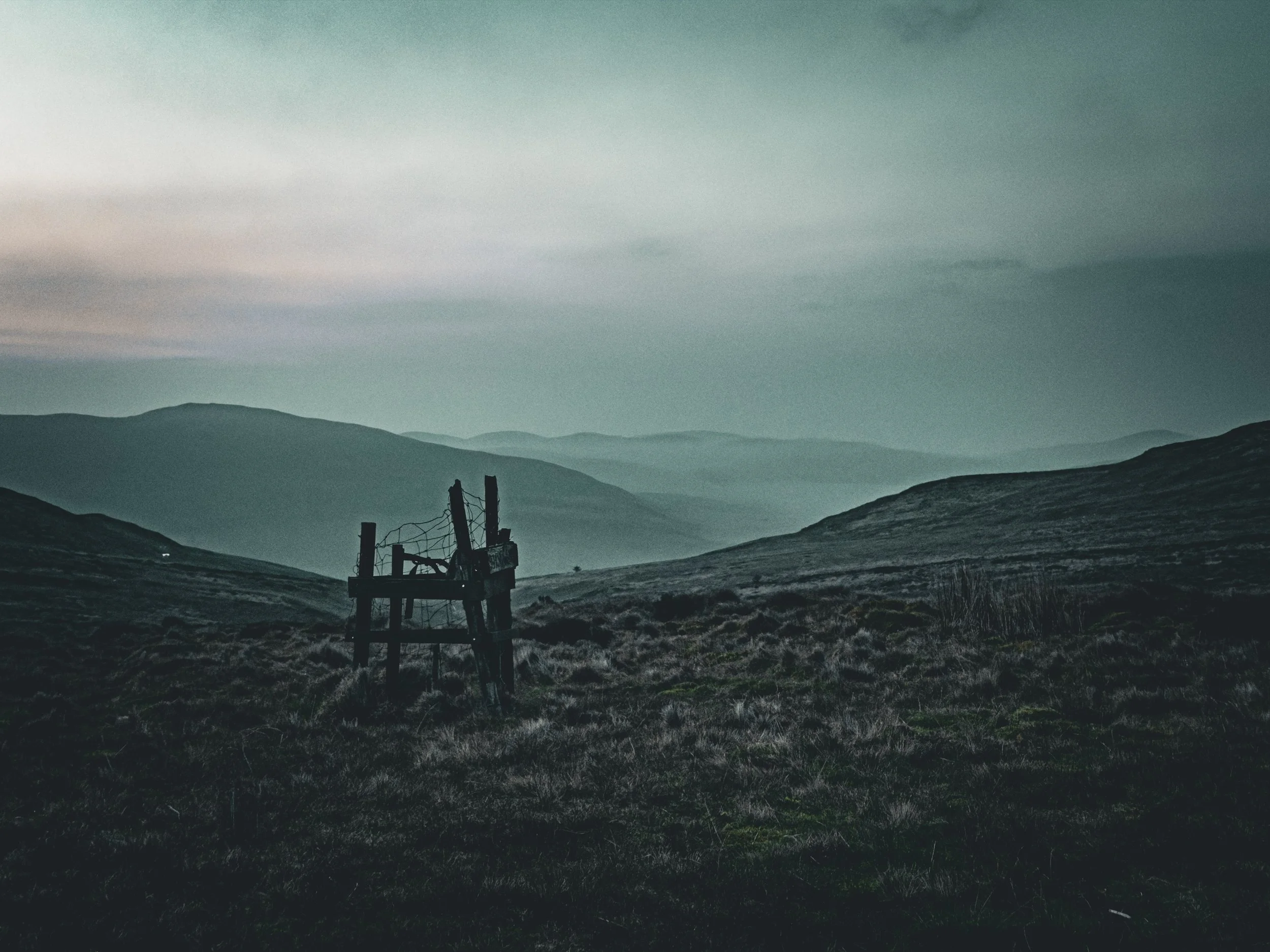 A desolate landscape with rolling hills and a broken wooden fence in the foreground under a cloudy sky.