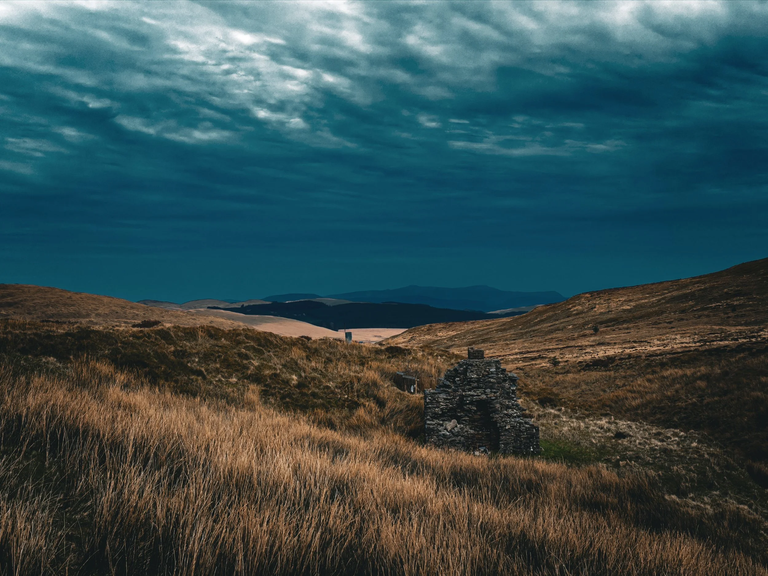 A rural landscape with rolling hills covered in tall, dry grass under a cloudy sky, and an old stone structure in the foreground.