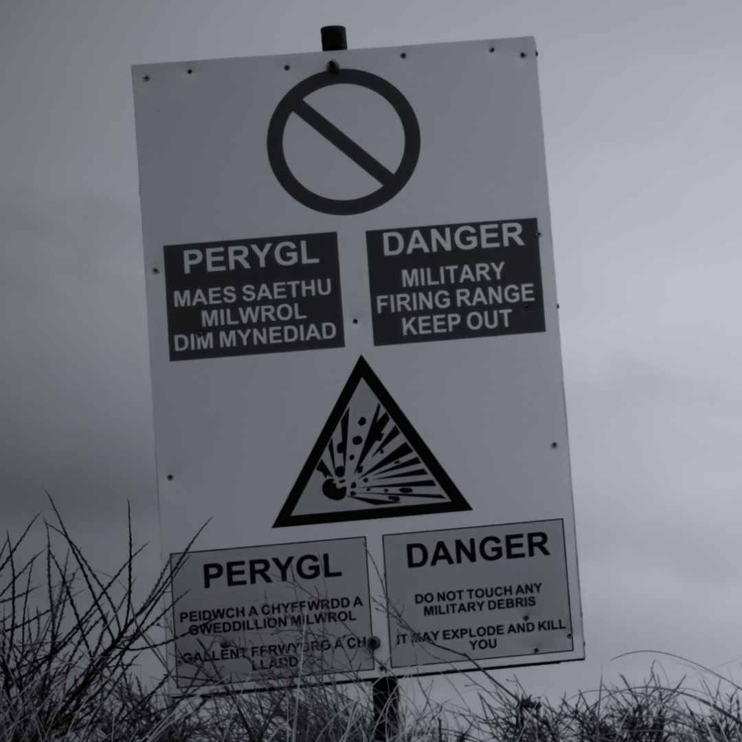 Sign warning of military firing range with danger symbols, no entry notices, and Welsh language warnings about military debris. The background shows an overcast sky and tall grass.
