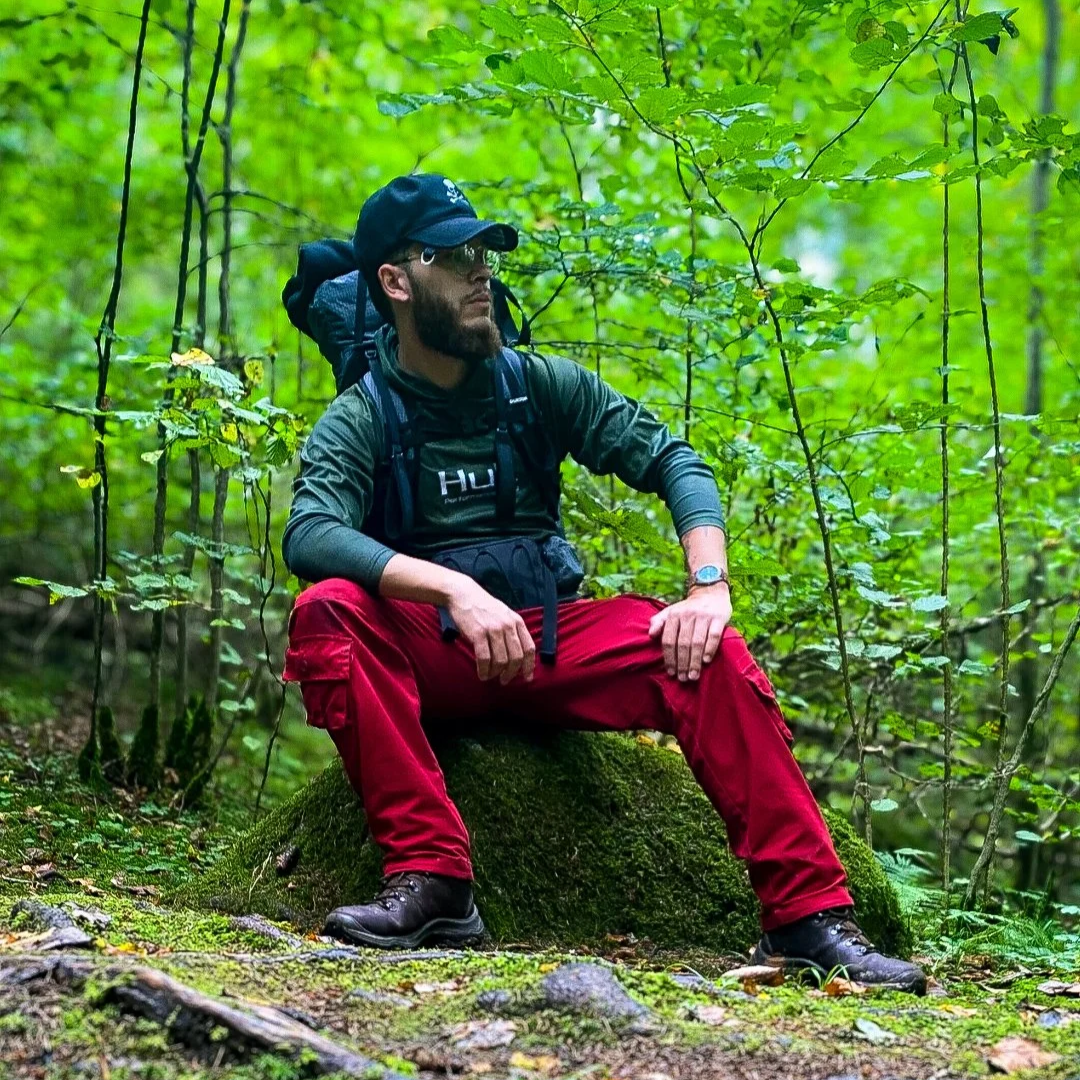 A man sitting on a mossy log in a lush green forest, wearing a black cap, sunglasses, a dark jacket, red pants, and hiking boots, with a backpack.