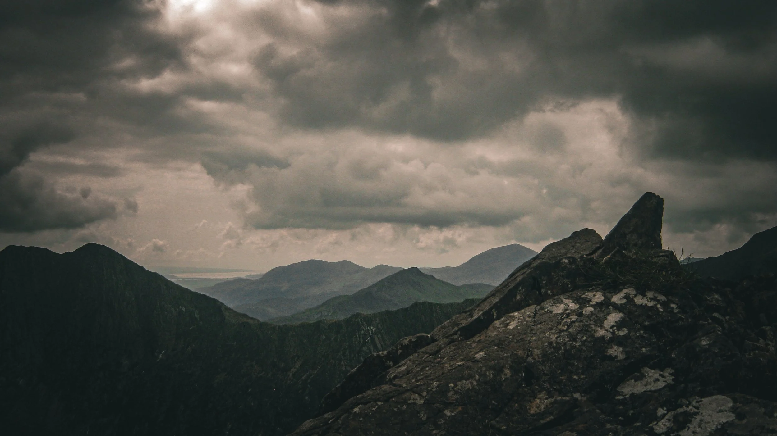 Mountain landscape under a cloudy sky with dark, rugged peaks in the foreground and distant mountains in the background.
