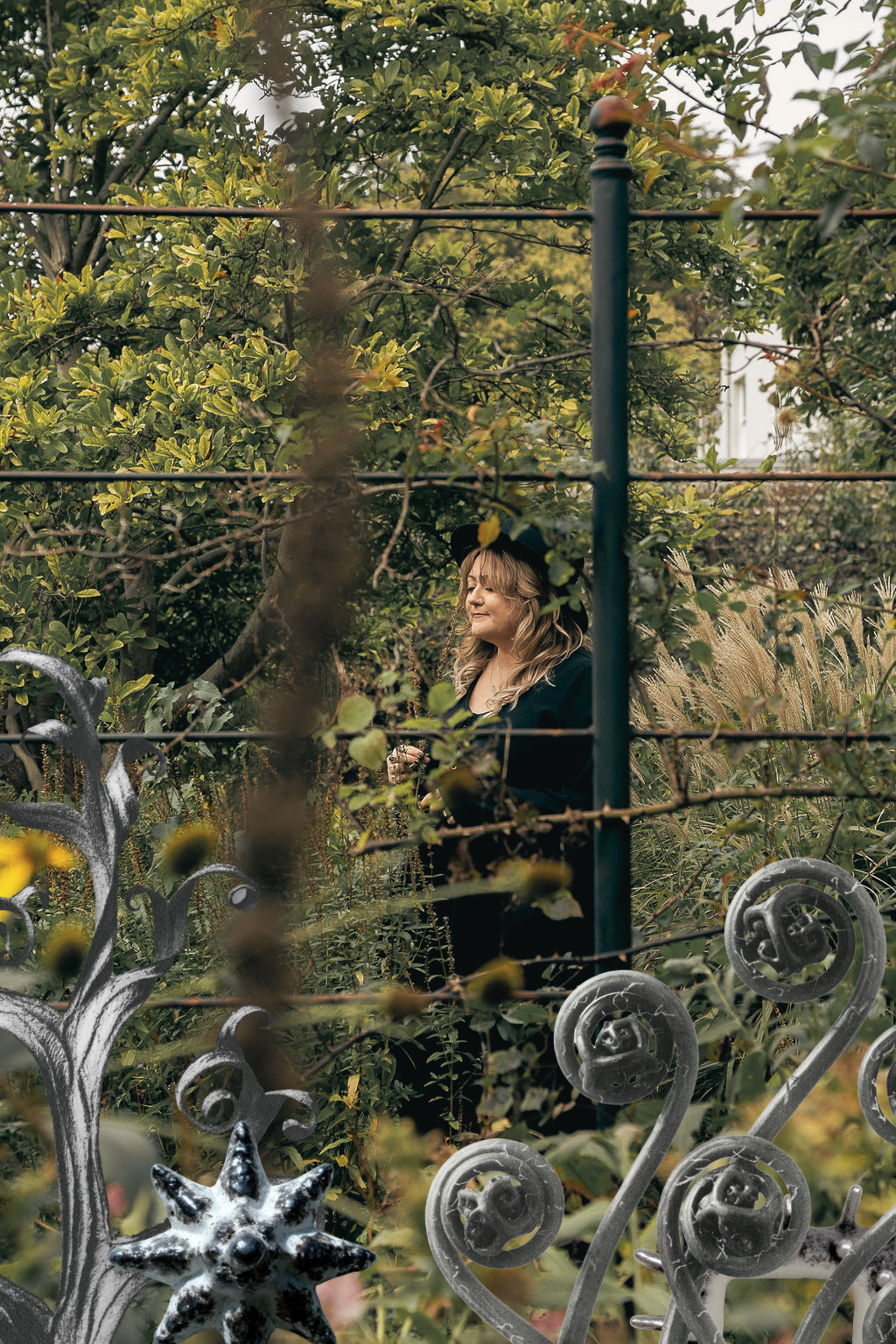 A woman with blonde hair and a black hat stands behind a garden fence, surrounded by green foliage and tall grasses, with her eyes closed and a peaceful expression.