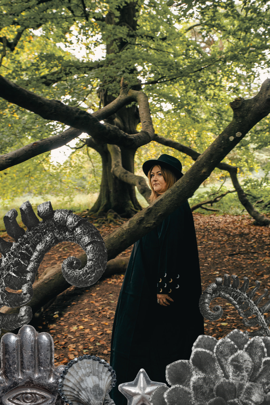 A woman standing in a forest with green leaves, wearing a black hat and black cloak decorated with white crescent moons, surrounded by black and silver decorative objects.