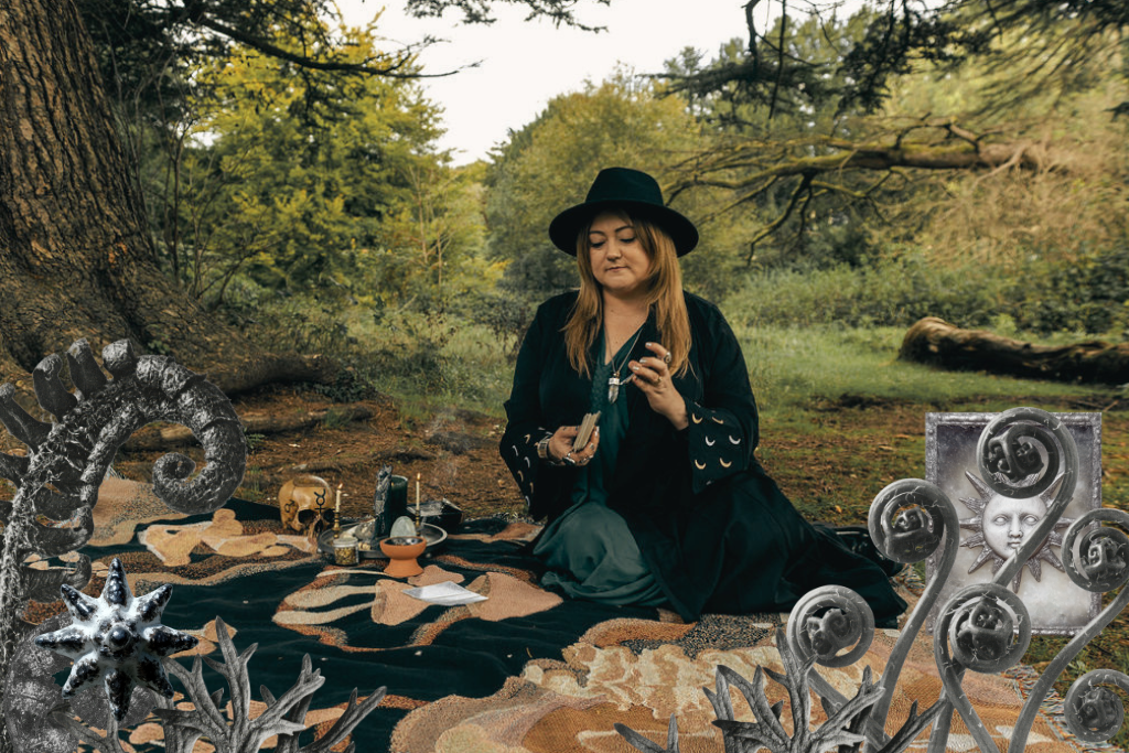 A woman dressed as a witch, wearing a black hat and cloak, sitting on a blanket in a forest clearing surrounded by mystical objects, with various fantastical decorations around her.