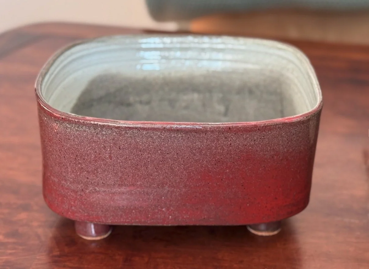 A small, square, red ceramic container with three small round feet, placed on a wooden surface.