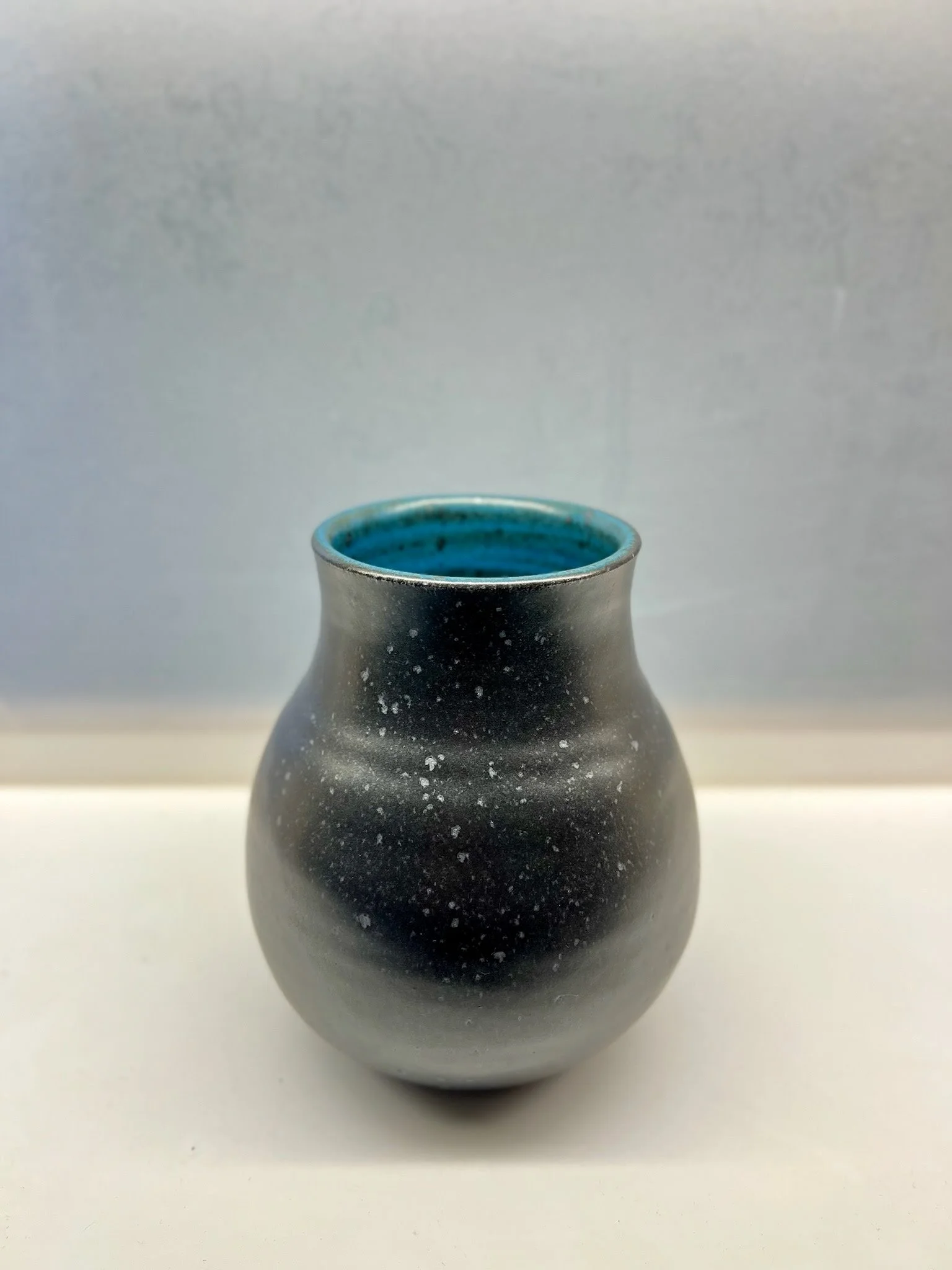 Black ceramic vase with blue interior, placed on white surface against a light background.