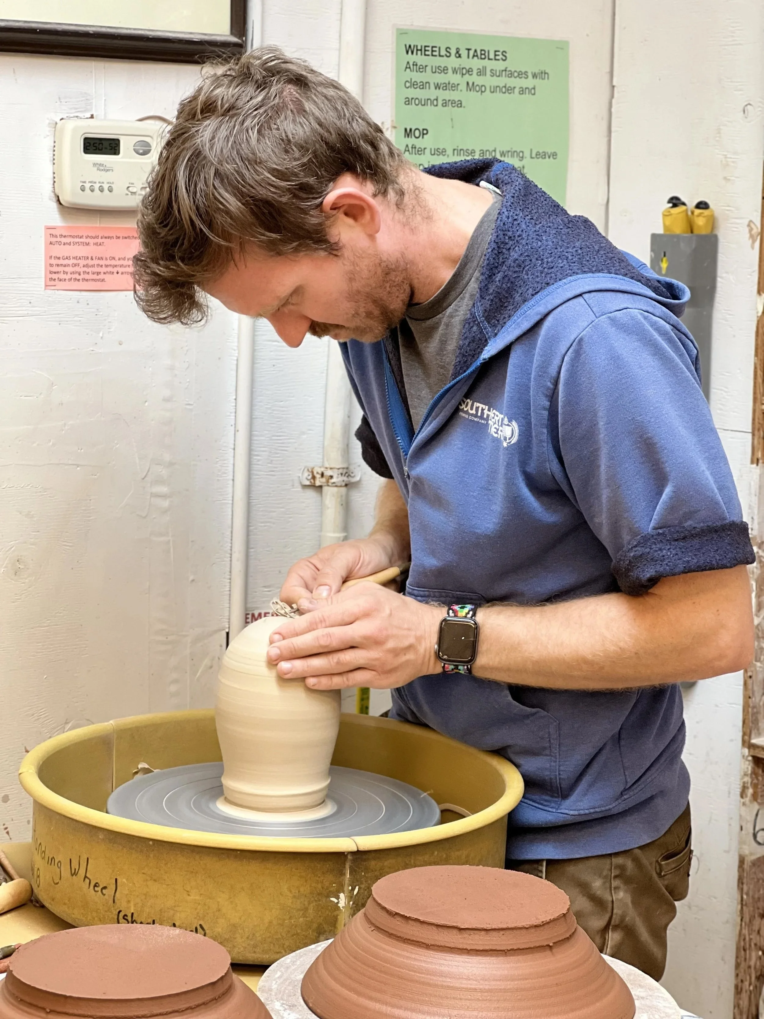 Zack Conquer trimming a ceramic vase on a pottery wheel in a pottery studio.