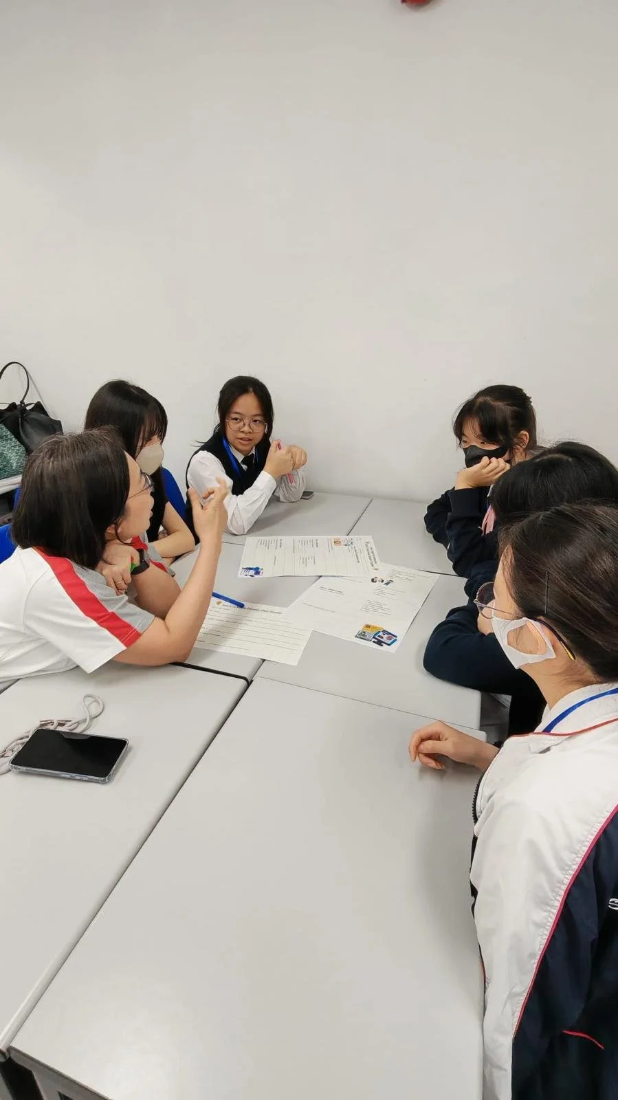 Six young girls wearing face masks and glasses sitting around a table engaged in a discussion with some papers and a smartphone on the table.