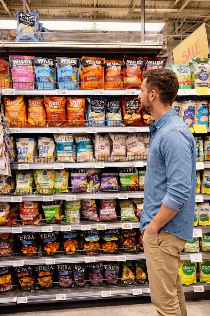 A man wearing a denim shirt and khaki pants shopping at a grocery store, standing in front of shelves filled with various snack bags including chips and protein snacks.
