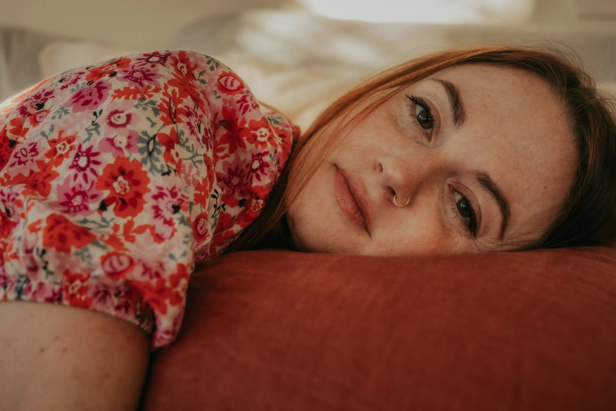 A woman with long red hair and freckles lying on her side on a bed, resting her head on a red pillow, wearing a floral-patterned shirt.
