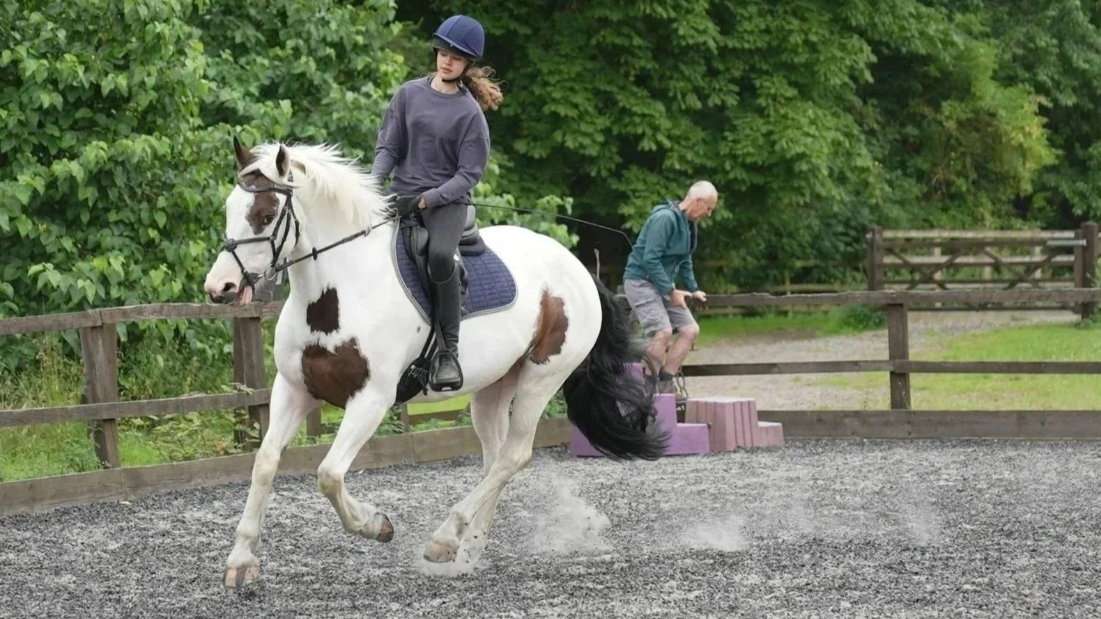 A girl riding a white horse with brown spots in an outdoor riding arena, with a man on a jump training exercise in the background.