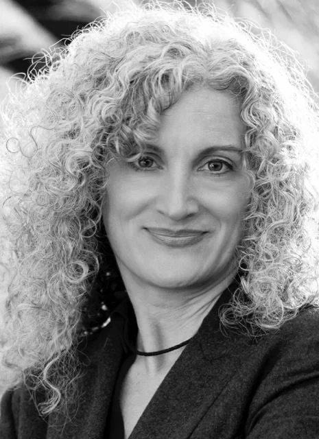 Black and white portrait of a woman with curly blonde hair, smiling, wearing a dark blazer and a choker necklace.