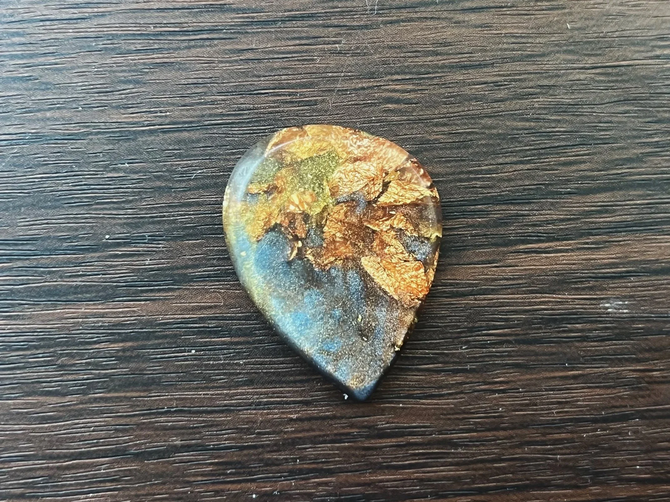 A semi-precious stone with a polished surface showing earthy colors, including green, brown, and gold, resting on a dark textured wooden surface.
