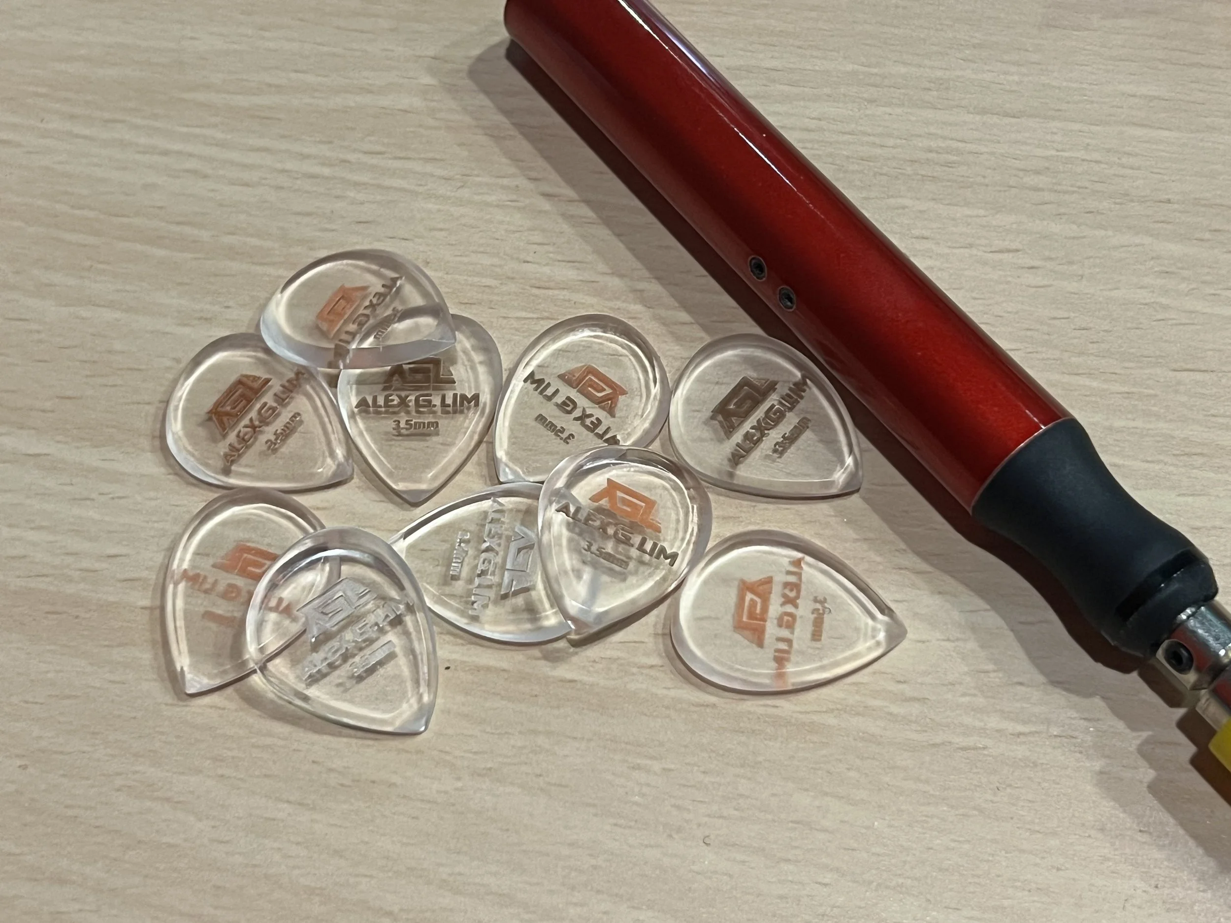 Red electric hair clipper with a black handle lying on a wooden surface, surrounded by multiple transparent guitar picks with the logo and text 'ALEXE HIM' and '35mm' printed on them.