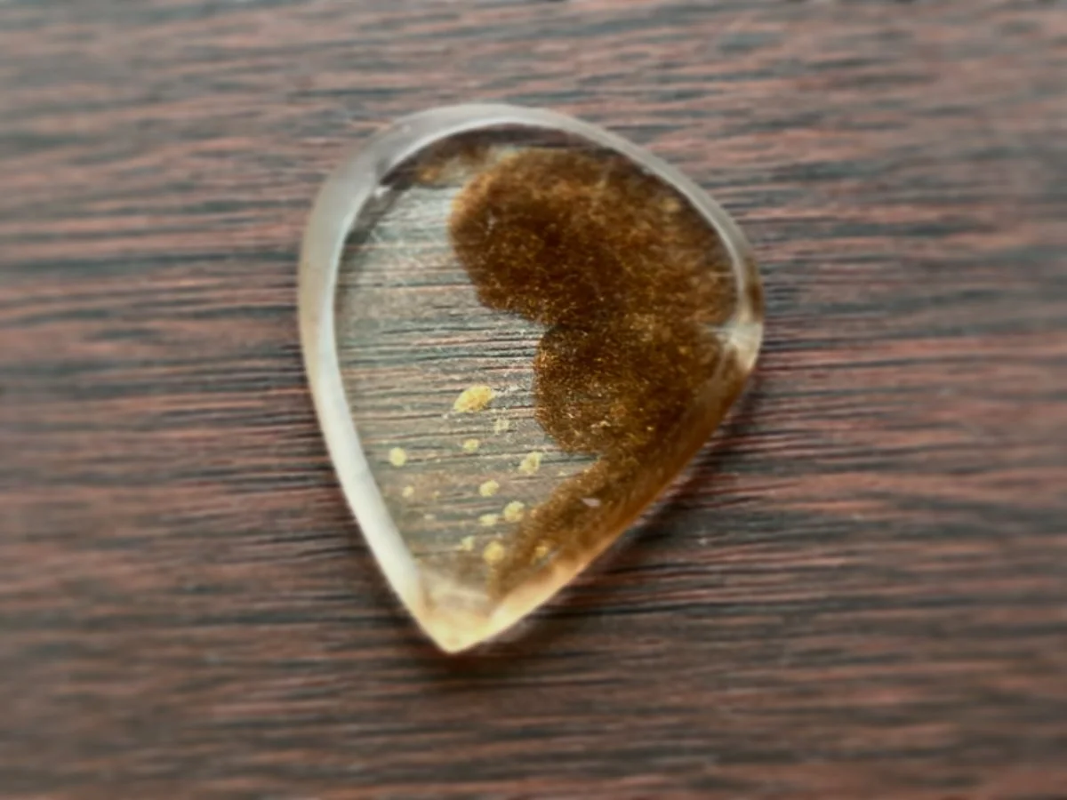 Close-up of a guitar pick with a clear edge and a brown, textured interior, placed on a wooden surface.