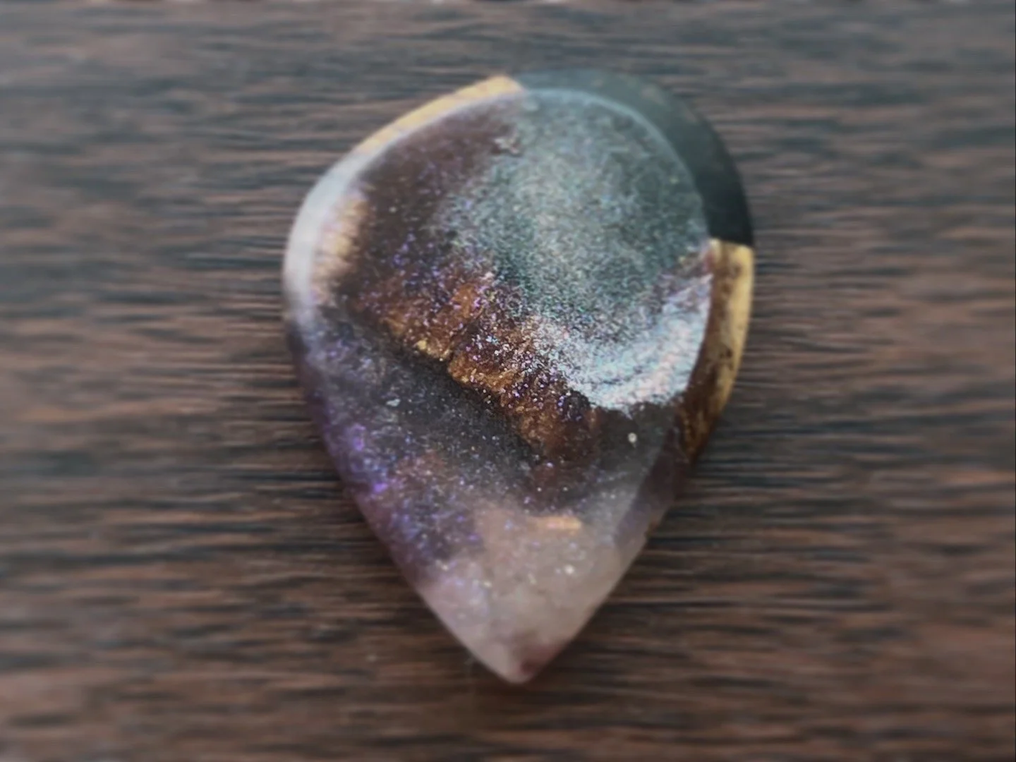 Close-up of a polished gemstone with a teardrop shape, displaying iridescent colors and a smooth surface, resting on a wooden surface.