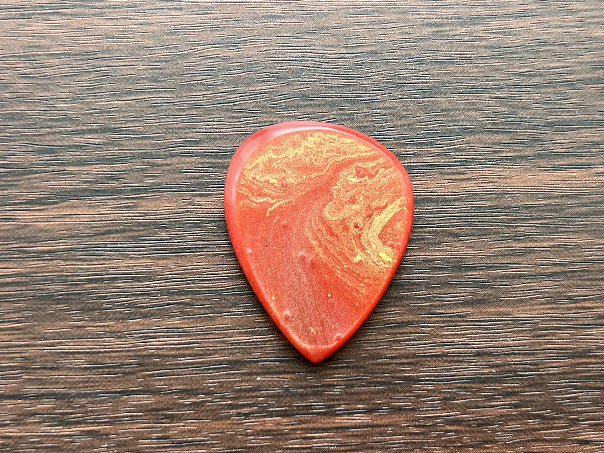 A red guitar pick with a marbled pattern lying on a wooden surface.
