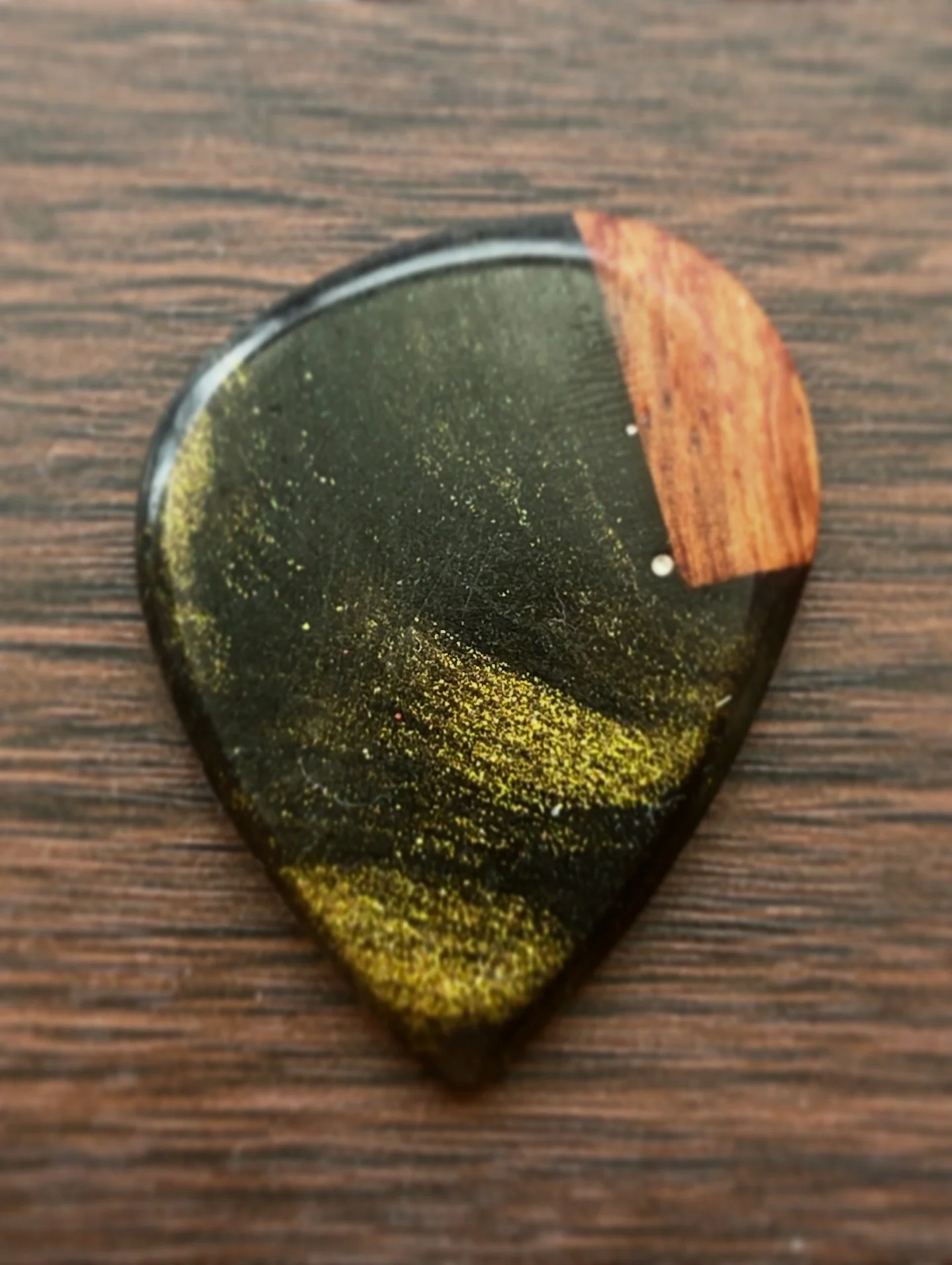 Close-up of a guitar pick with a black and gold speckled design and a small orange-brown wood inlay on a wooden surface.