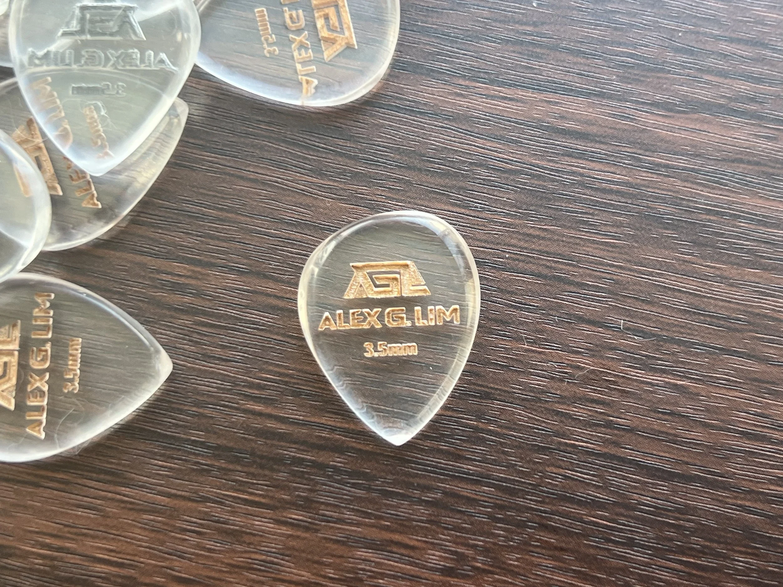 Close-up of transparent guitar picks with gold inscription 'ALEX G LIM 3.5 mm' on a wooden surface.