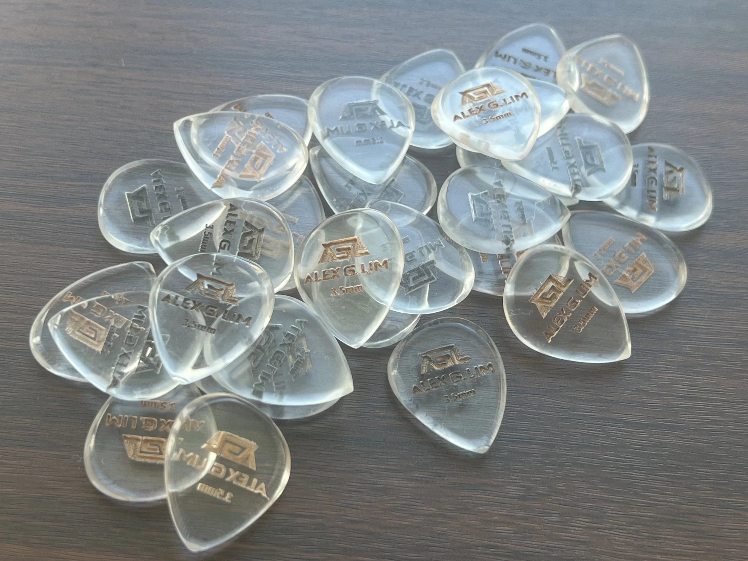 A pile of transparent guitar picks with gold and black text, some overlapping, on a dark wooden surface.