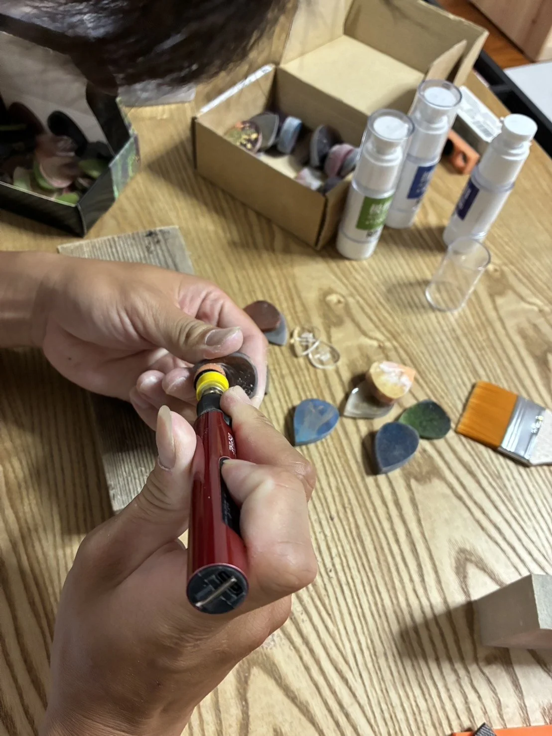 Person carving a piece of stone or mineral with a rotary tool on a wooden table with various stones, paints, and art supplies scattered around.