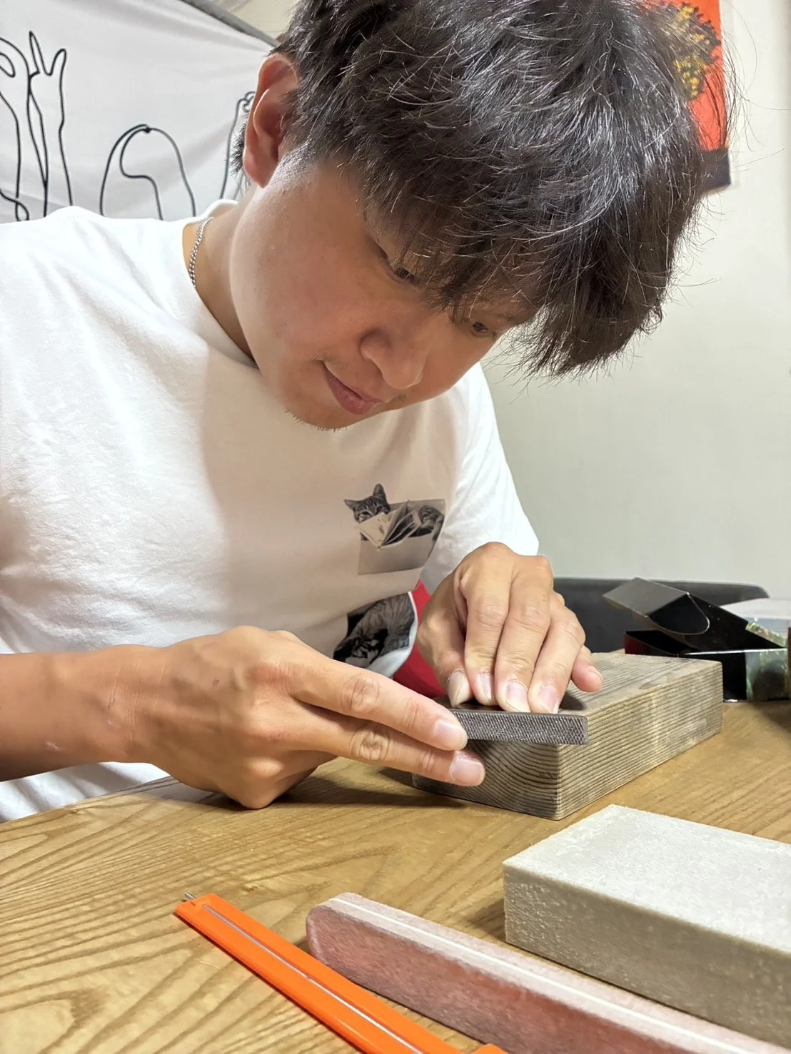 A man shaping a piece of stone or ceramic with a file, sitting at a wooden table with tile materials and tools around him.