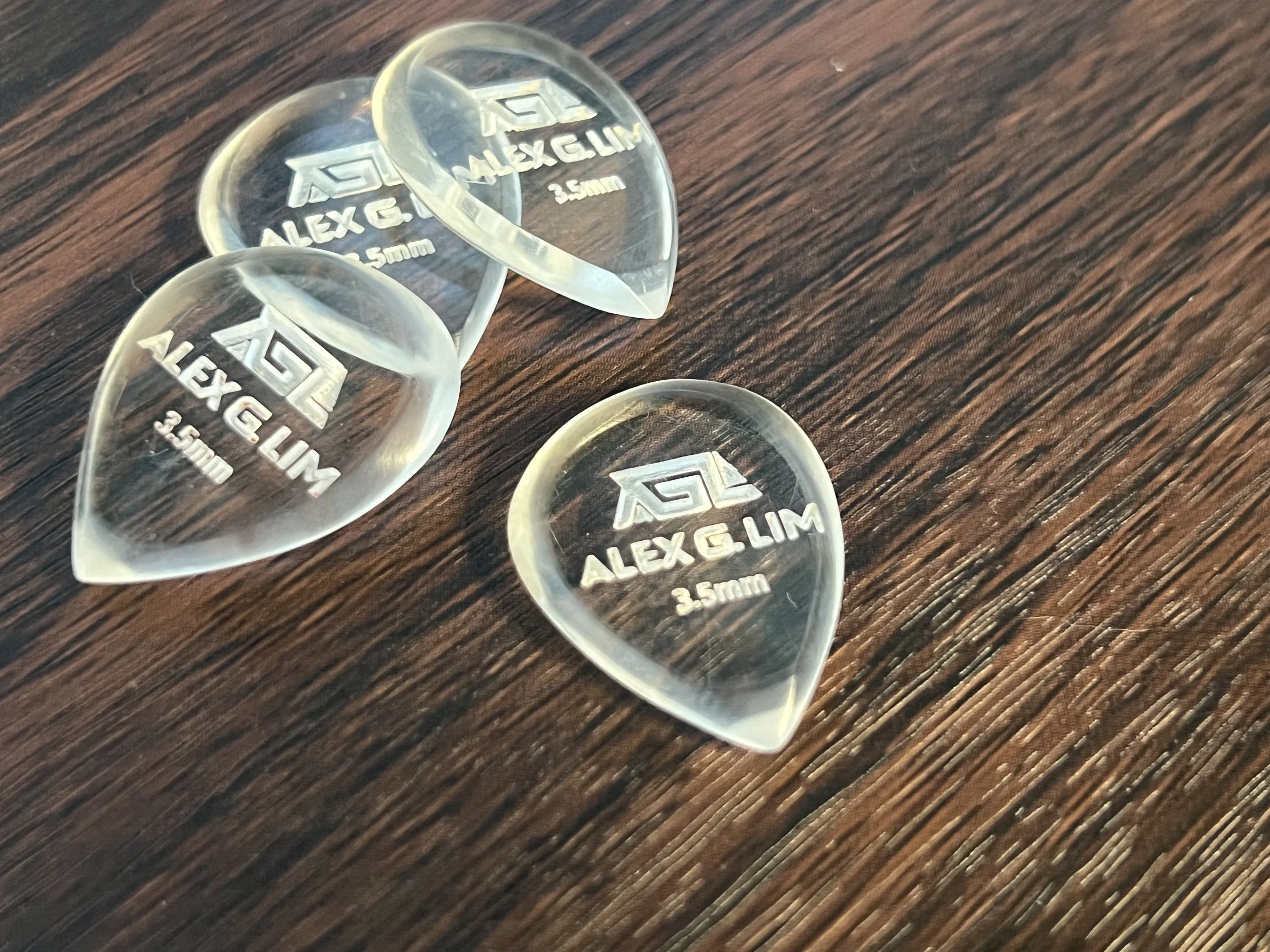 Three transparent plastic guitar picks on a wooden surface, engraved with 'ALEX G. ALEXELIM 3.5mm' and a logo.