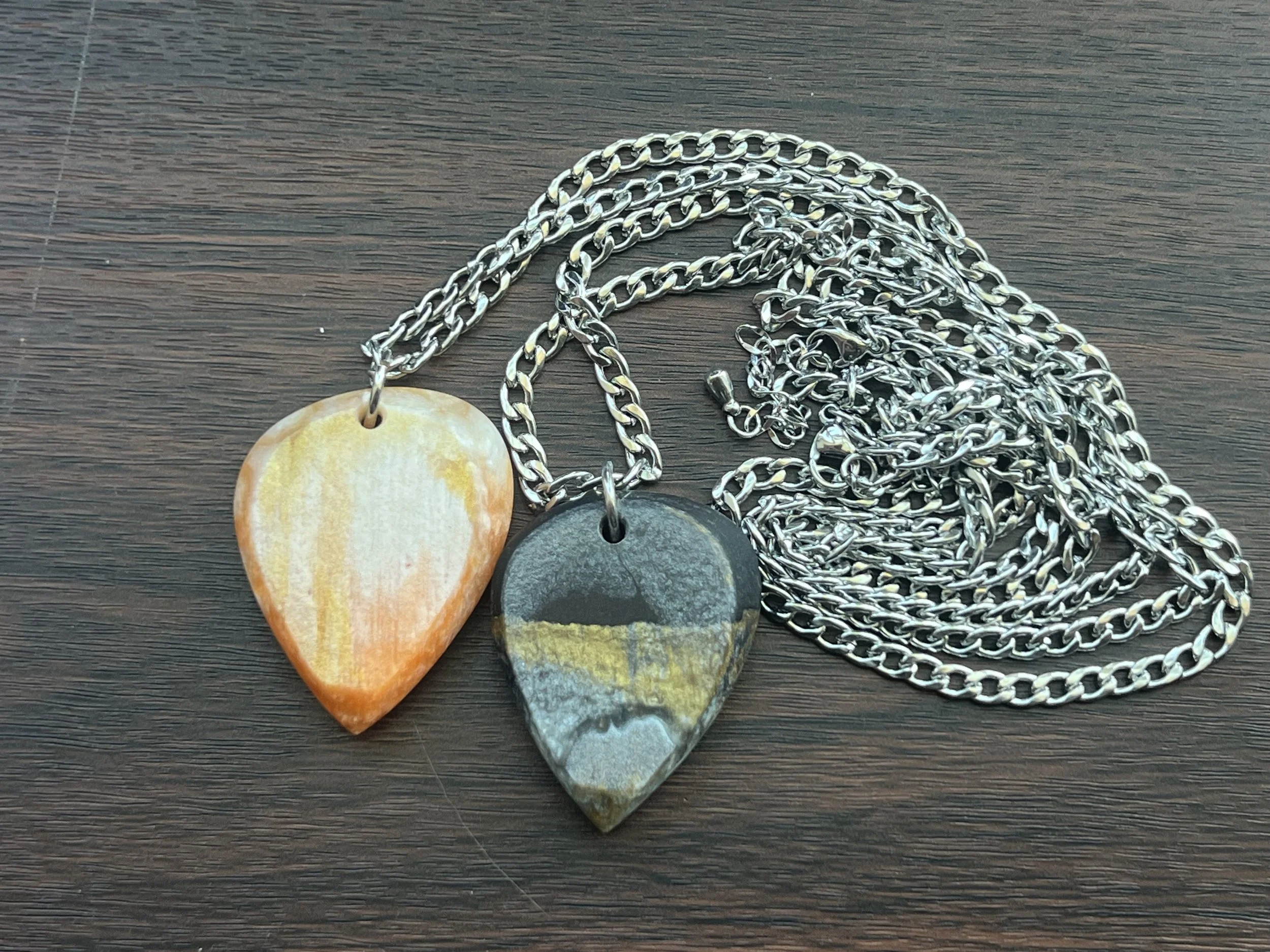 Three necklaces with pendant charms placed on a wooden surface. The charms include a beige and orange shell, a dark stone, and a multicolored stone with yellow, black, and white hues.
