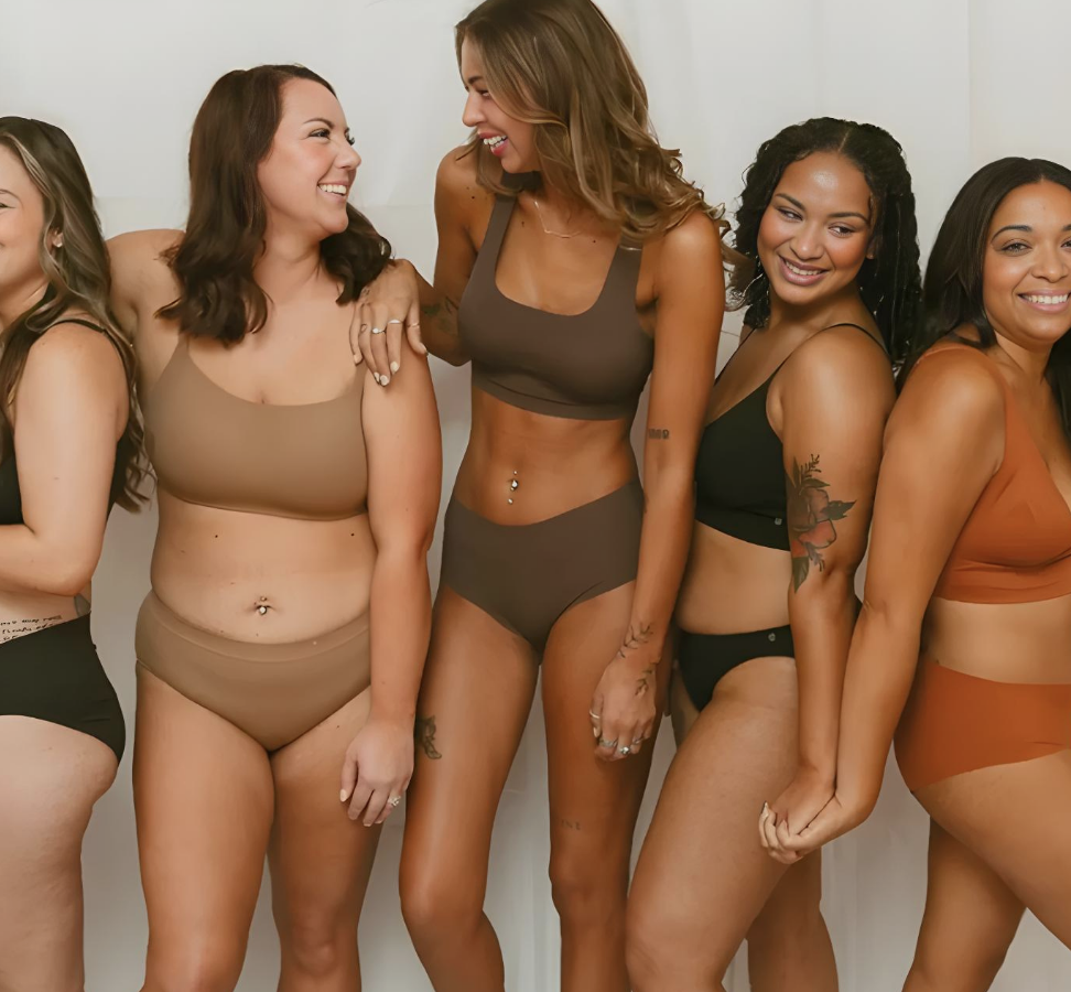 Group of five diverse women in underwear laughing and smiling near a white wall.