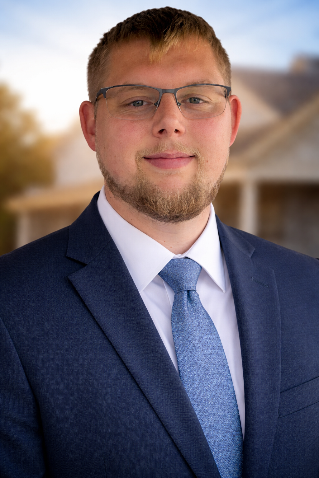 Portrait of a man wearing glasses, a navy suit, a white shirt, and a blue tie, standing outdoors with a blurred background of trees and a house.