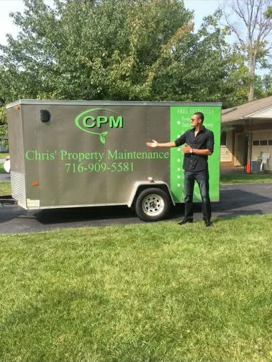 A man in black clothes standing in front of a green and gray property maintenance trailer, holding a clipboard. The trailer has the company name 'Chris' Property Maintenance' with a phone number, and the logo 'CPM' in green.
