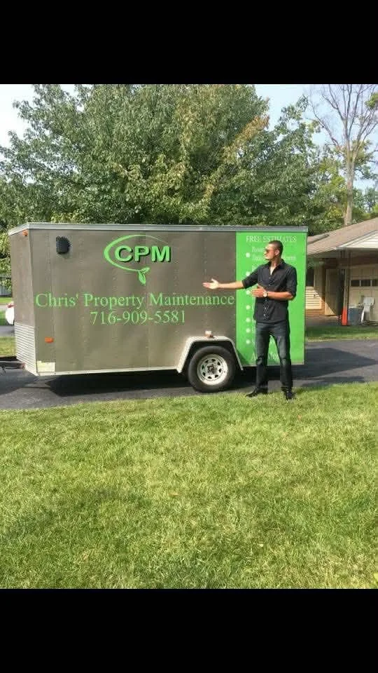 A man standing next to a mobile property maintenance trailer, pointing at the trailer, with a grassy yard, trees, and a house in the background.
