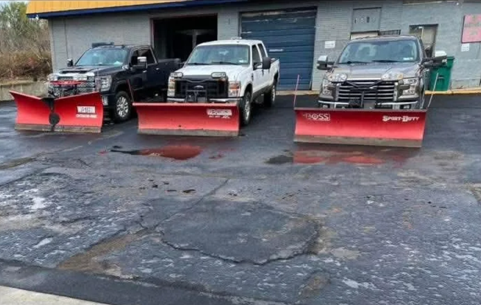Three trucks parked in front of a gray building with a garage door, each equipped with a large red snow plow attached to the front.