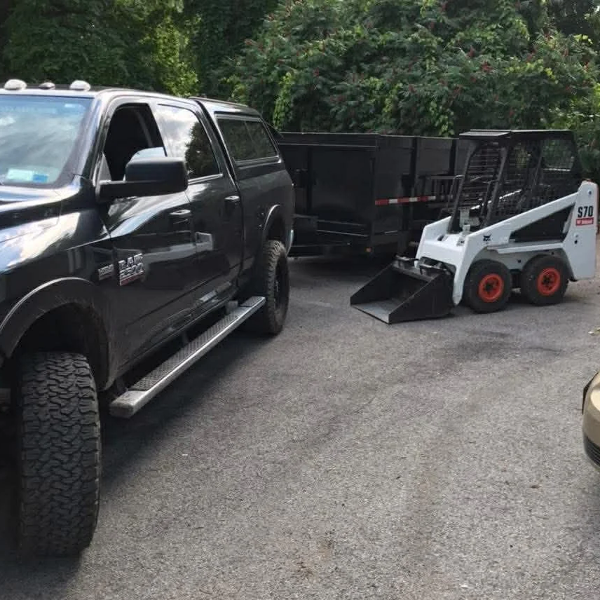 CPM Inc.'s truck with a black trailer and a small skid-steer loader, parked on the side of a road with trees in the background.