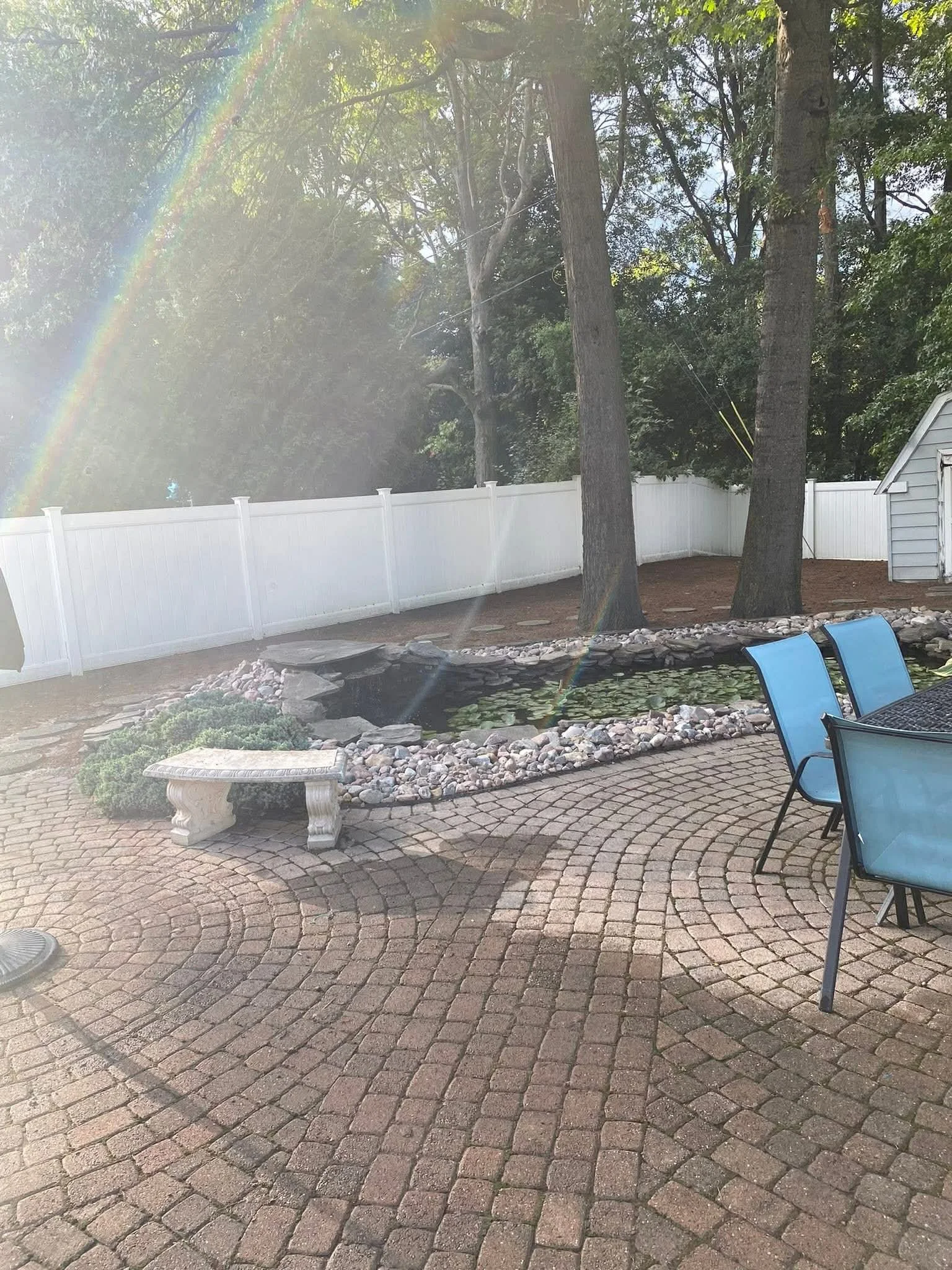 A backyard patio with brick pavers, a small pond with lily pads, a white fence, tall trees, a stone bench, blue chairs, and a small shed in the background, with a rainbow and sunlight streaming through the trees.