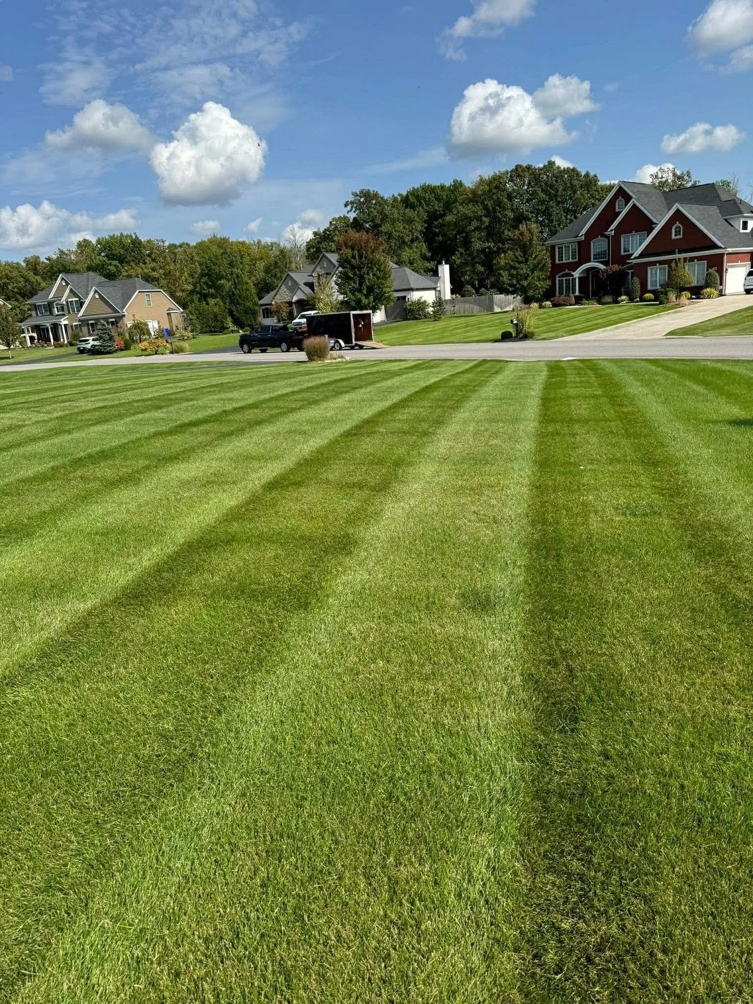 Well-manicured green lawn with alternating light and dark green stripes, residential houses in the background under a bright blue sky with scattered white clouds.
