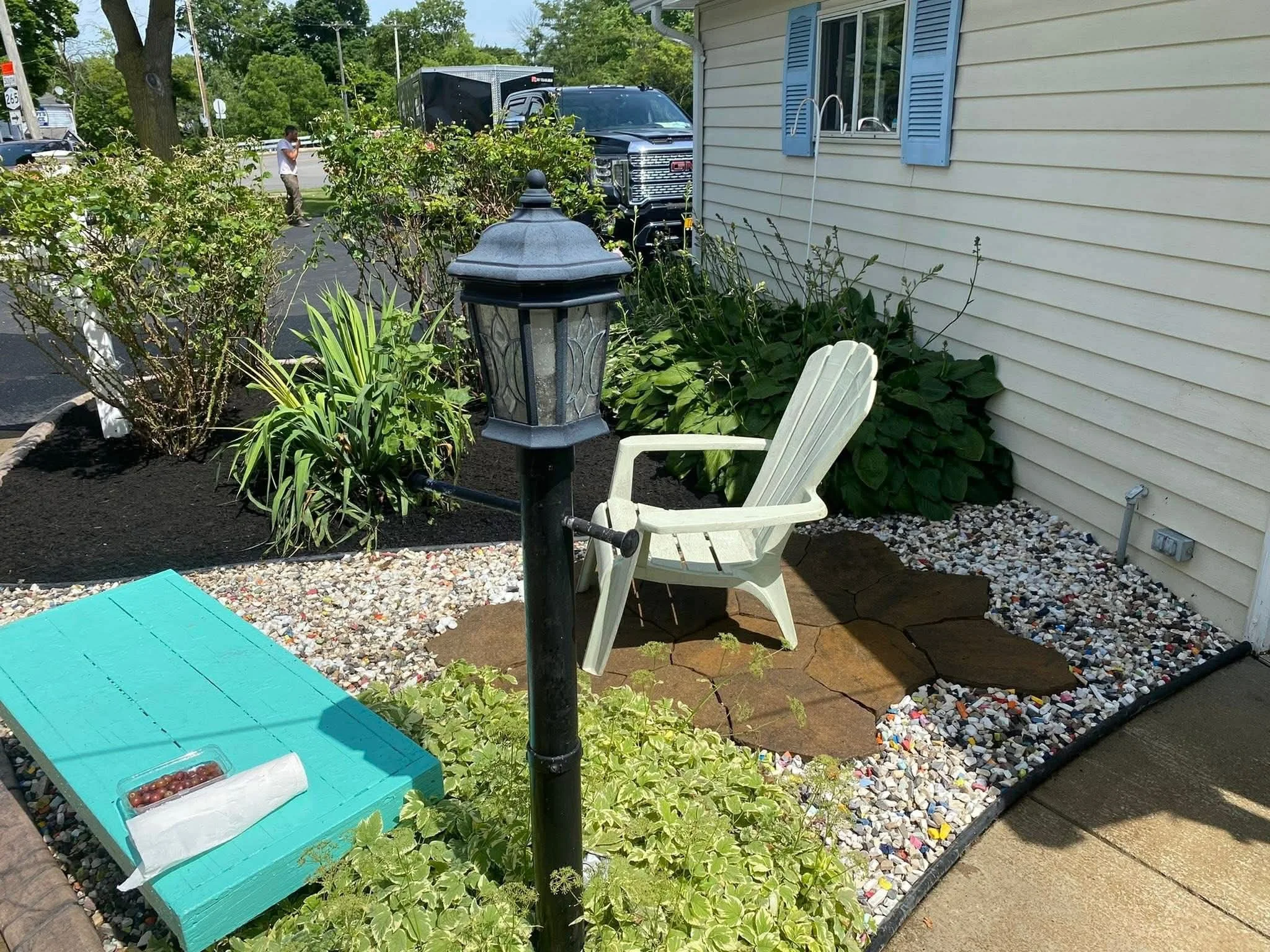 A small outdoor sitting area with a white plastic chair, a black lamp post, a blue wooden box with grapes and paper towel on top, surrounded by garden plants and crushed stones, next to a house with beige siding and blue shutters.