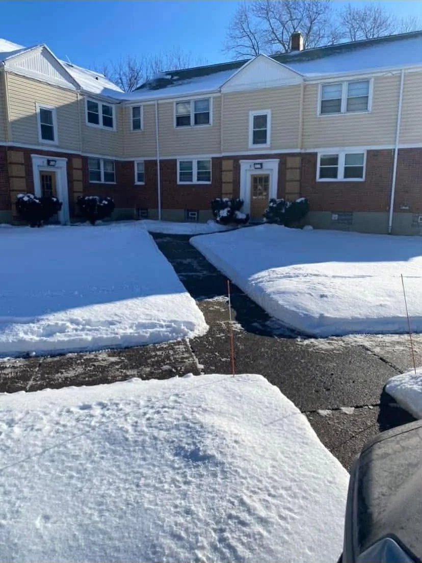 A residential street with a snow-covered sidewalk, driveway, and lawn in front of a two-story apartment complex. The apartment building has beige siding and brick on the lower level, with white-framed windows and doors. The sky is clear and blue, and there are a few leafless trees in the background.