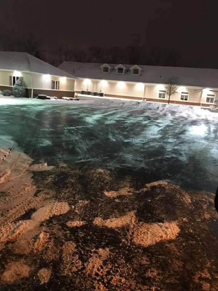 Snow-covered parking lot with tire tracks and footprints, illuminated building in the background at night.