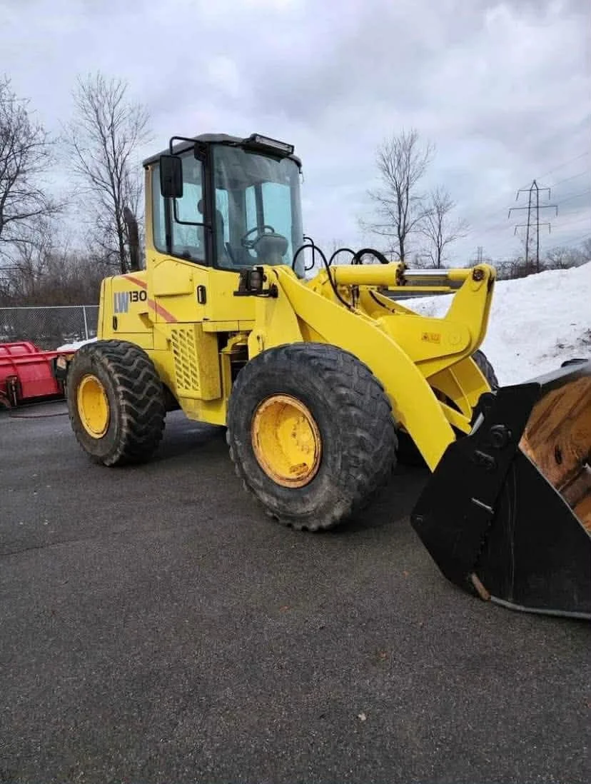 Yellow front loader with a snow plow attachment parked on an asphalt surface near snow, trees, and power lines under a cloudy sky.