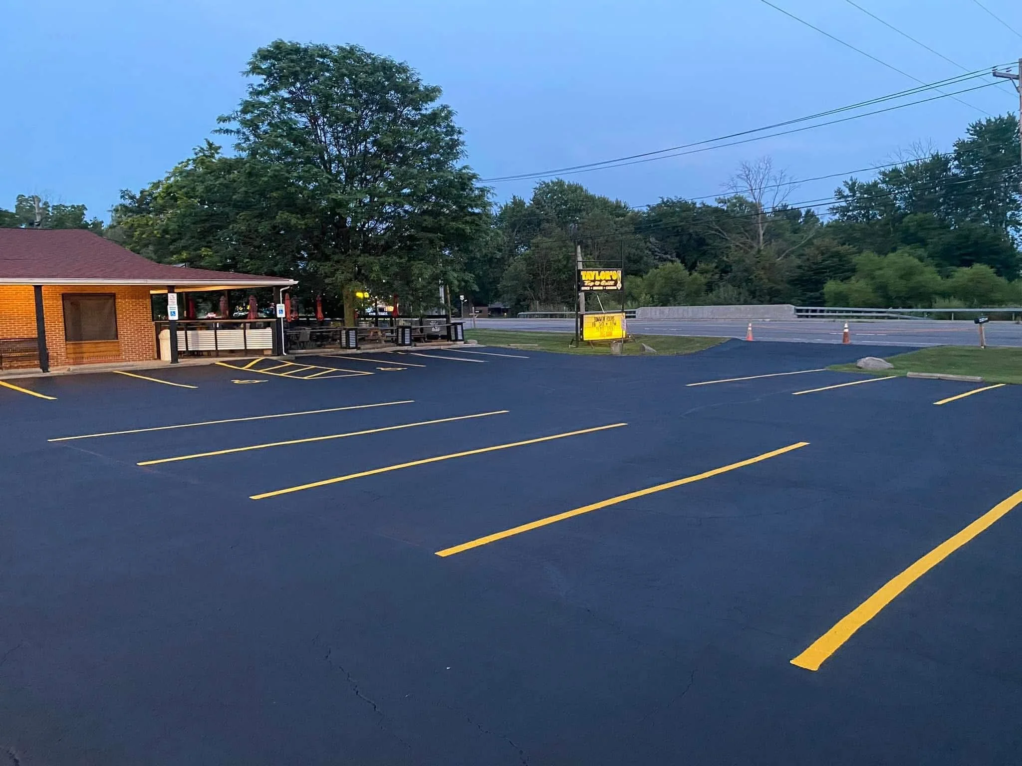 Empty parking lot outside a brick restaurant with a patio, with yellow parking space lines, a large tree, a street with traffic cones, and a yellow and black signboard in the background.