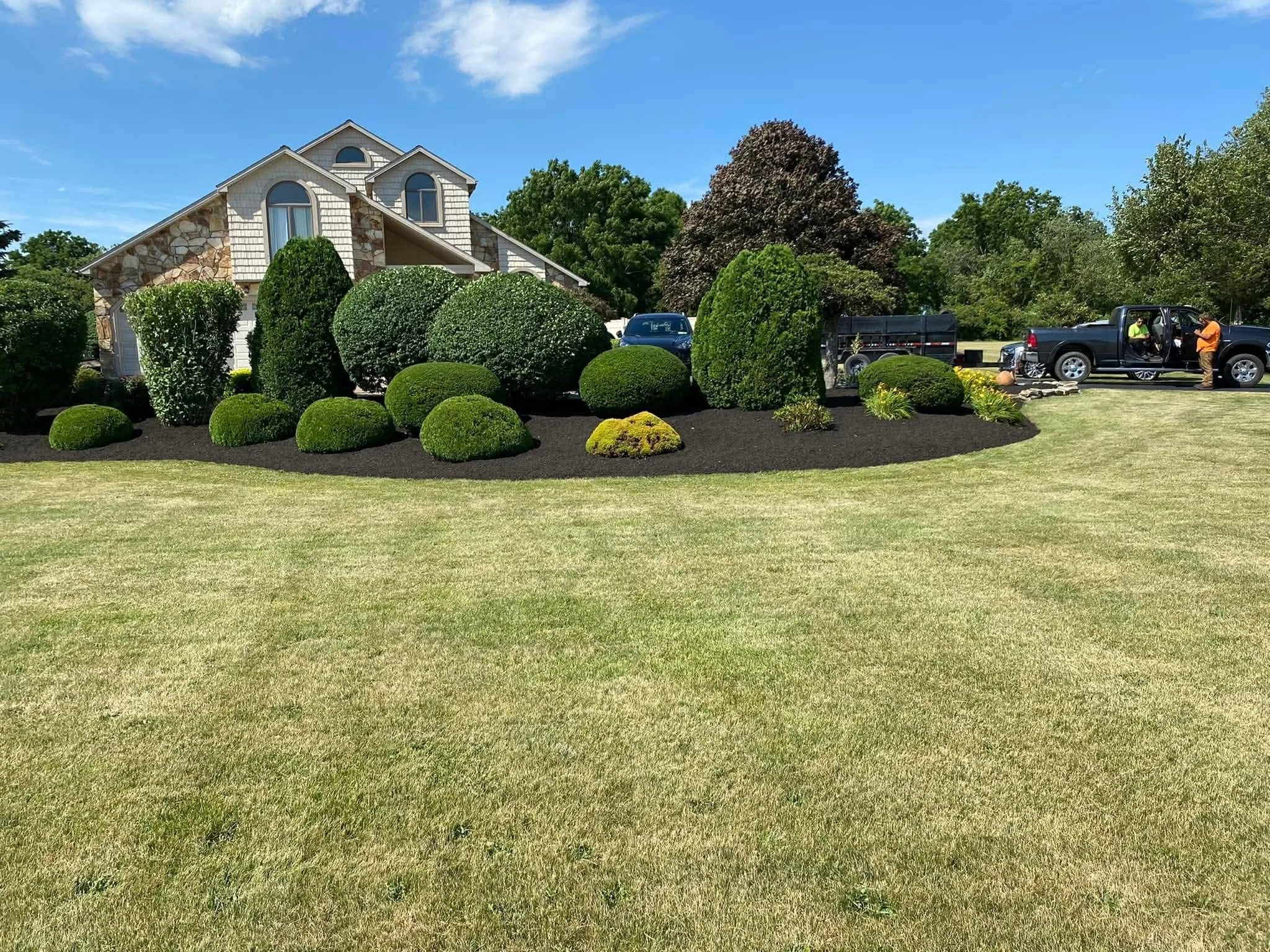 A well-maintained front yard with neatly trimmed bushes and a large house in the background, with two trucks and two workers near the trucks.