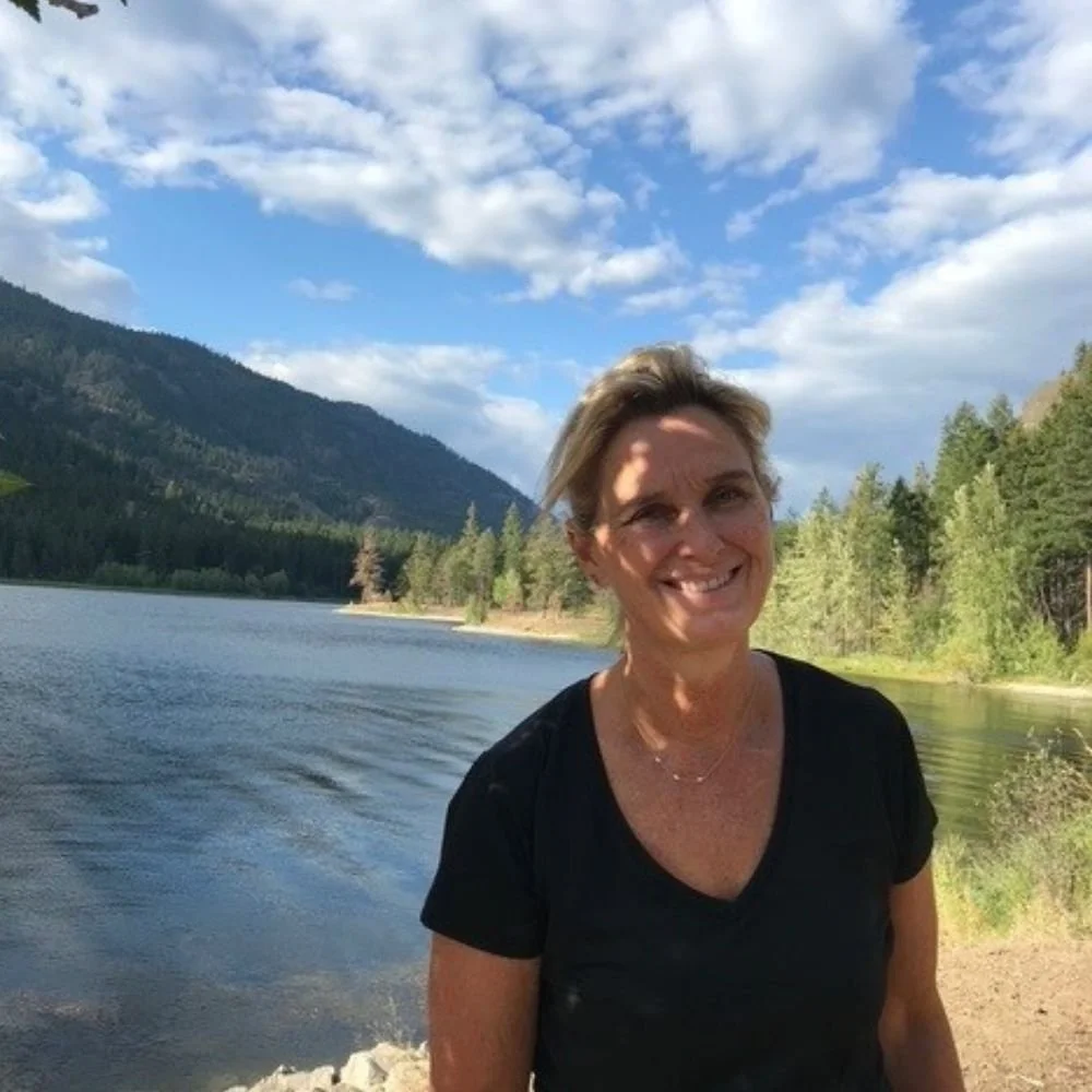 A smiling woman stands on a rocky shore next to a lake with a mountain and trees in the background under a partly cloudy sky.