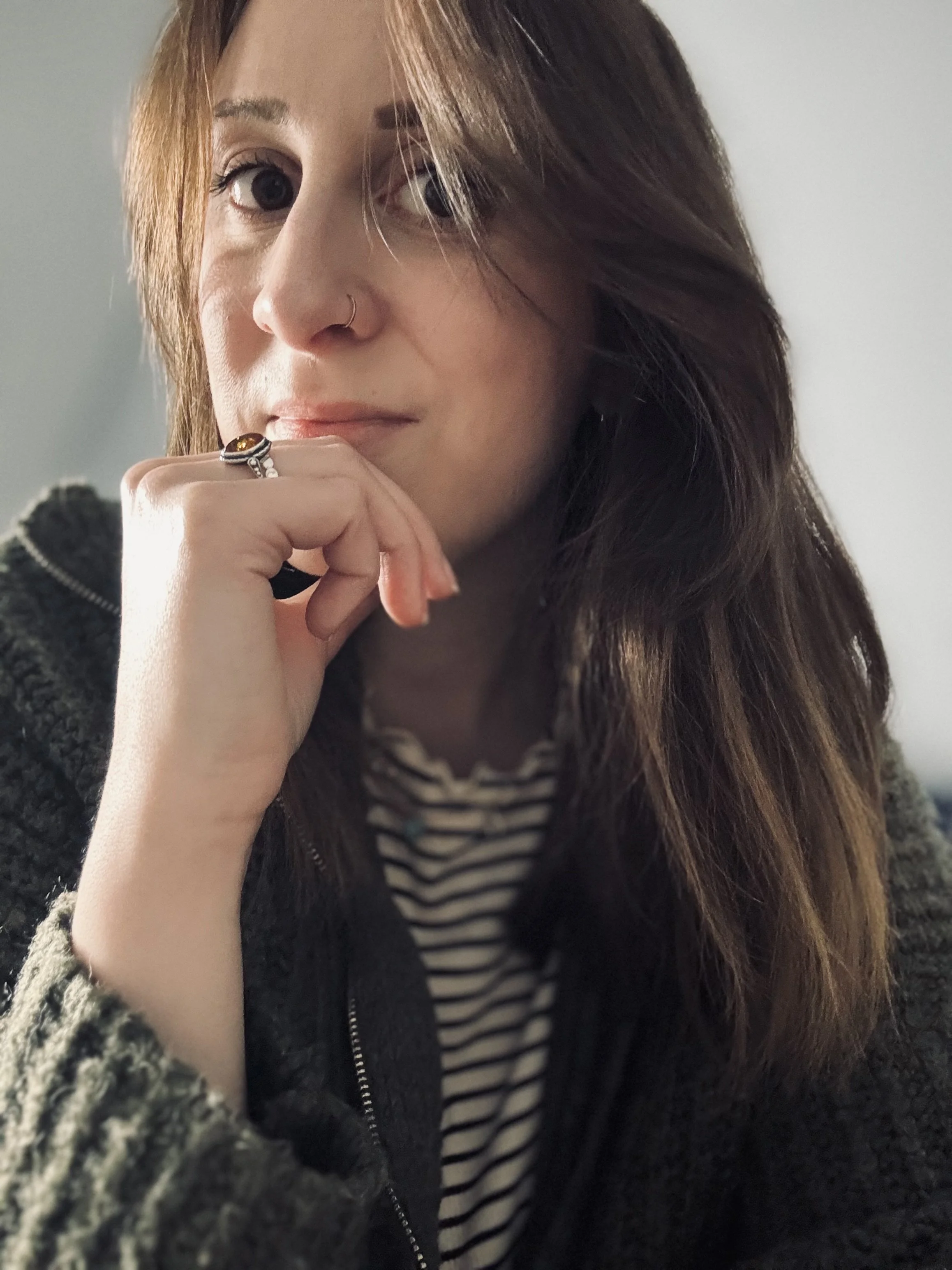 Portrait d'une femme aux cheveux longs bruns, portant une veste en laine sombre, une chemise rayée, et un anneau avec un pendentif orange sur l'annulaire de sa main droite, posée sous son menton. Elle a un piercing au nez et regarde vers l'objectif, dans un intérieur lumineux.
