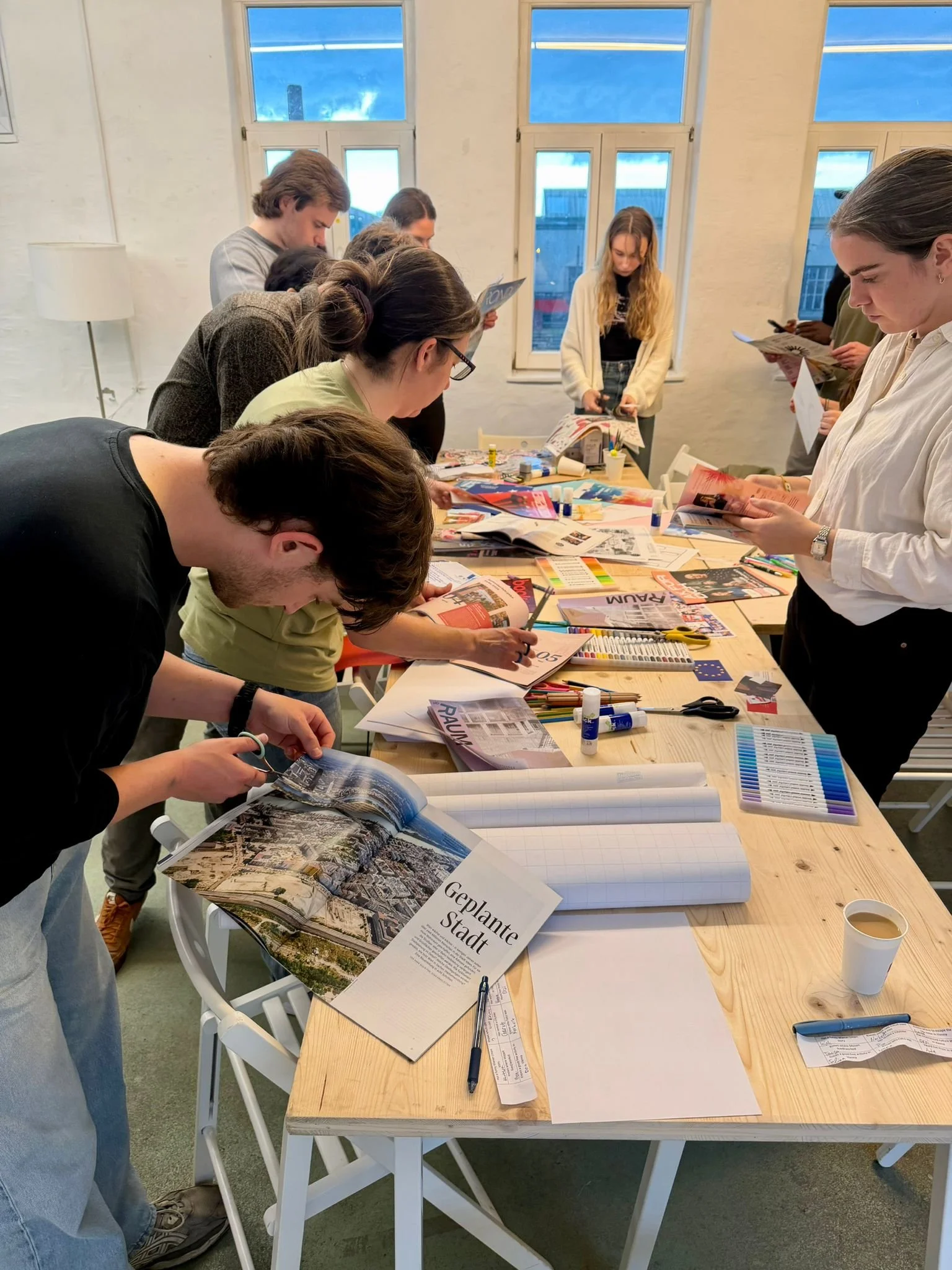 Group of people gathered around a large wooden table working on projects involving magazines, papers, and art supplies. The table has various tools like scissors, glue, and pens. The setting appears to be a bright room with large windows.