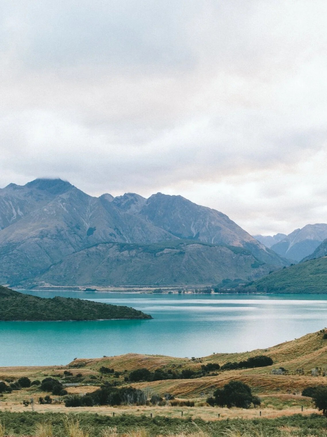 Scenic view of a lake surrounded by mountains with grassy fields and trees in the foreground under a cloudy sky.