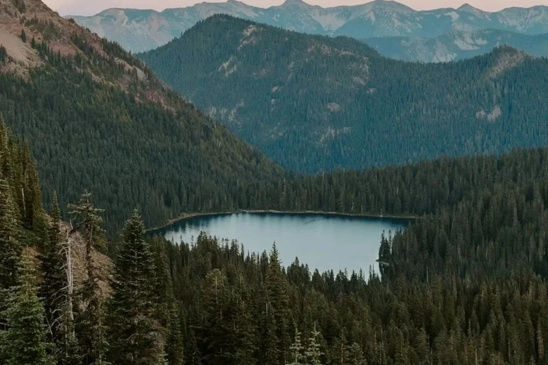 A mountain lake surrounded by dense evergreen forest under a partly cloudy sky.