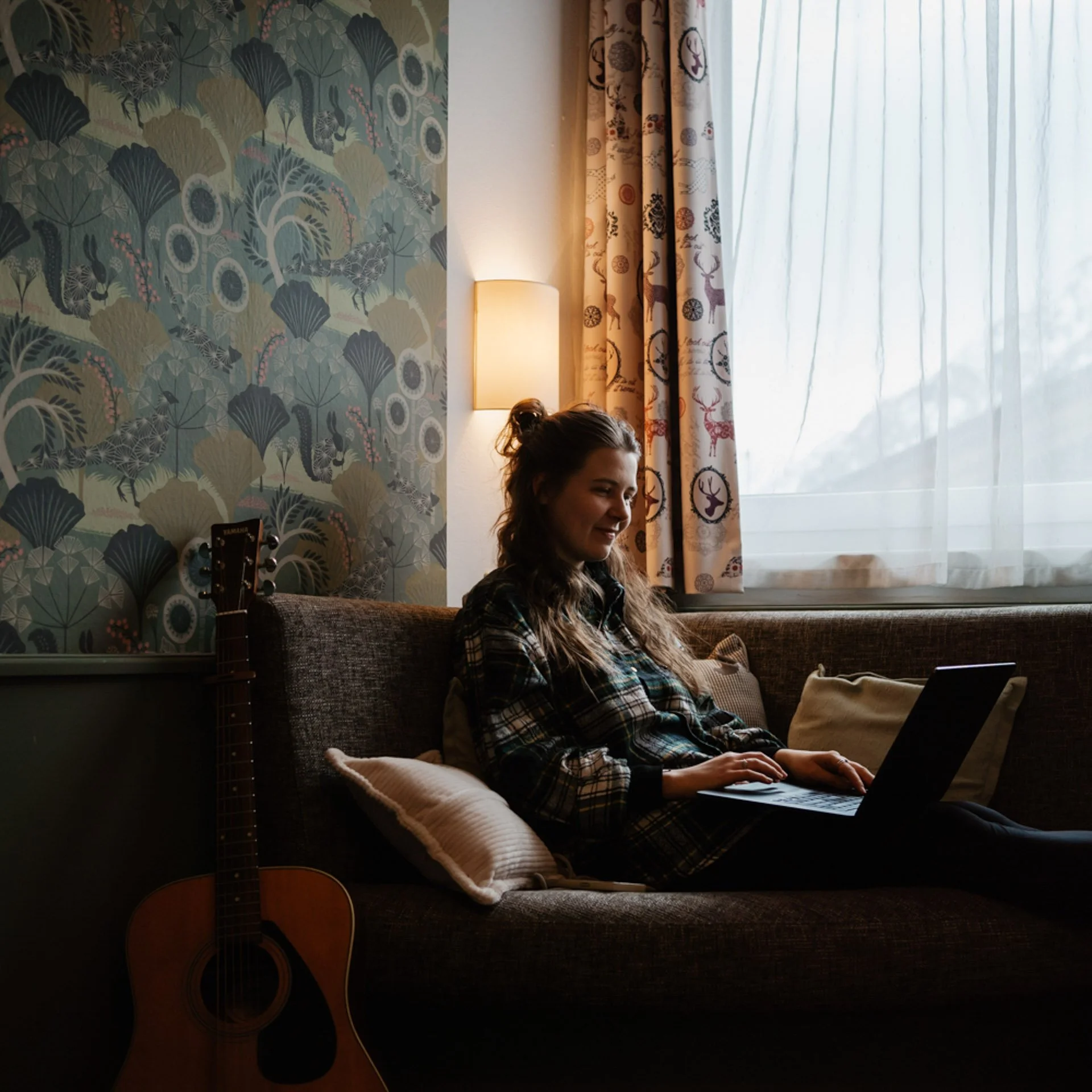 Junge Frau sitzt entspannt auf einem Sofa, arbeitet an einem Laptop, im Hintergrund beige Vorhänge mit Hirschmotiven und eine Wand mit blätterförmigem Muster, neben ihr eine Akustikgitarre.