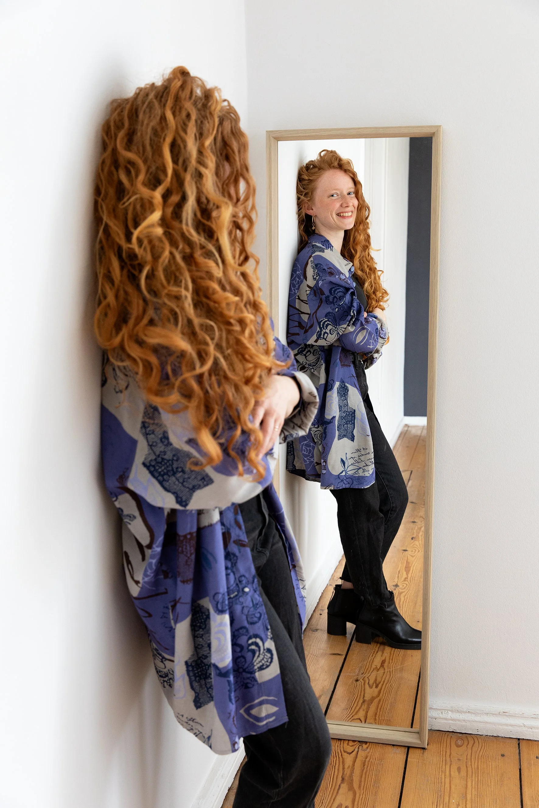 Eine Frau mit lockigem roten Haar in einer blauen, gemusterten Bluse steht vor einem Vollspiegelschrank auf Holzparkett, lächelt, trägt schwarze Hose und Stiefeletten, im Hintergrund eine weiße Wand.